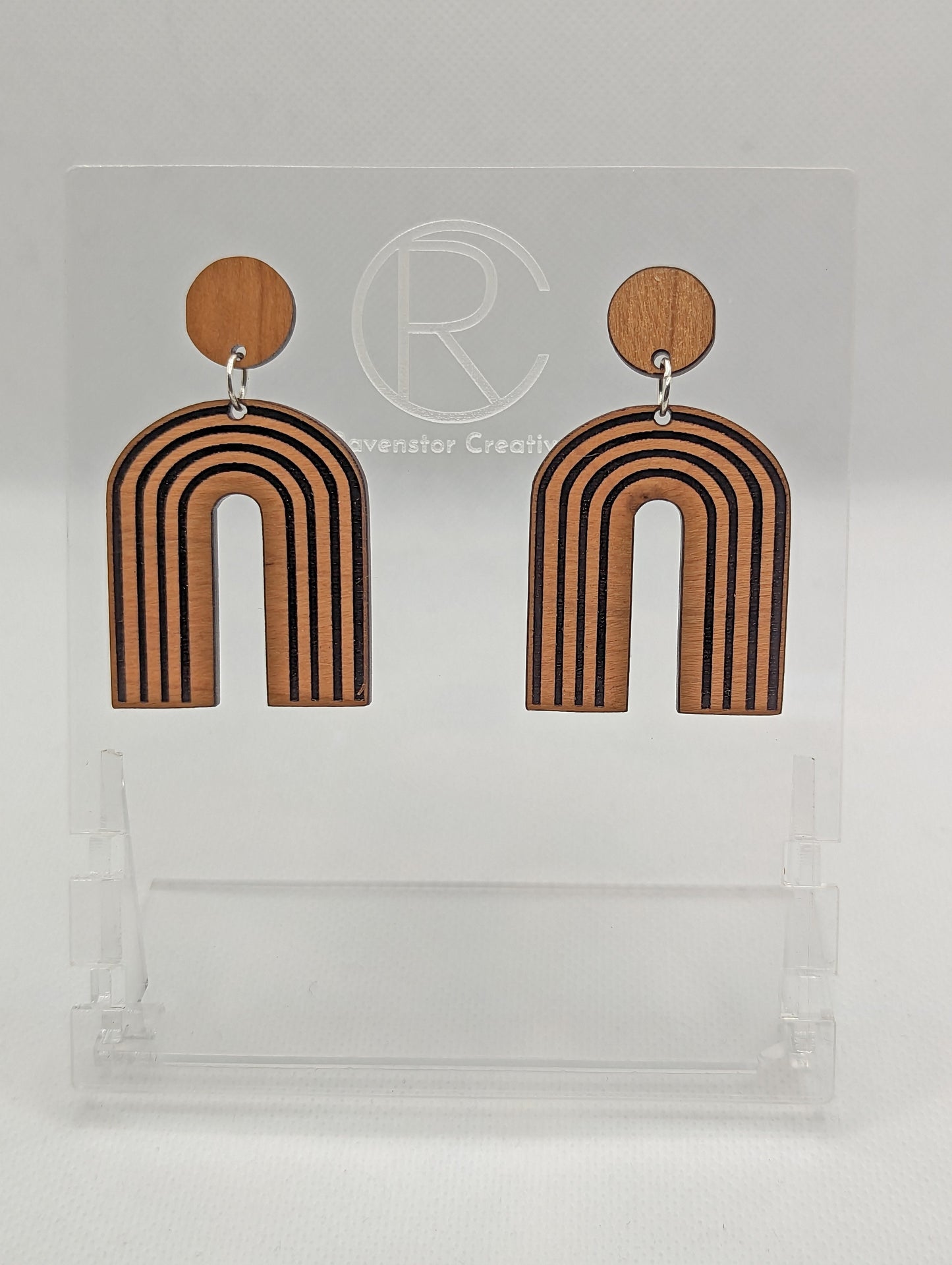 A pair of arch earrings dangling under a circle with stud fixings. Engraved are lines like a wooden rainbow 80s retro style. They are made from Cherry wood. They are against a white background on a clear stand with the Ravenstor Creative logo on it.