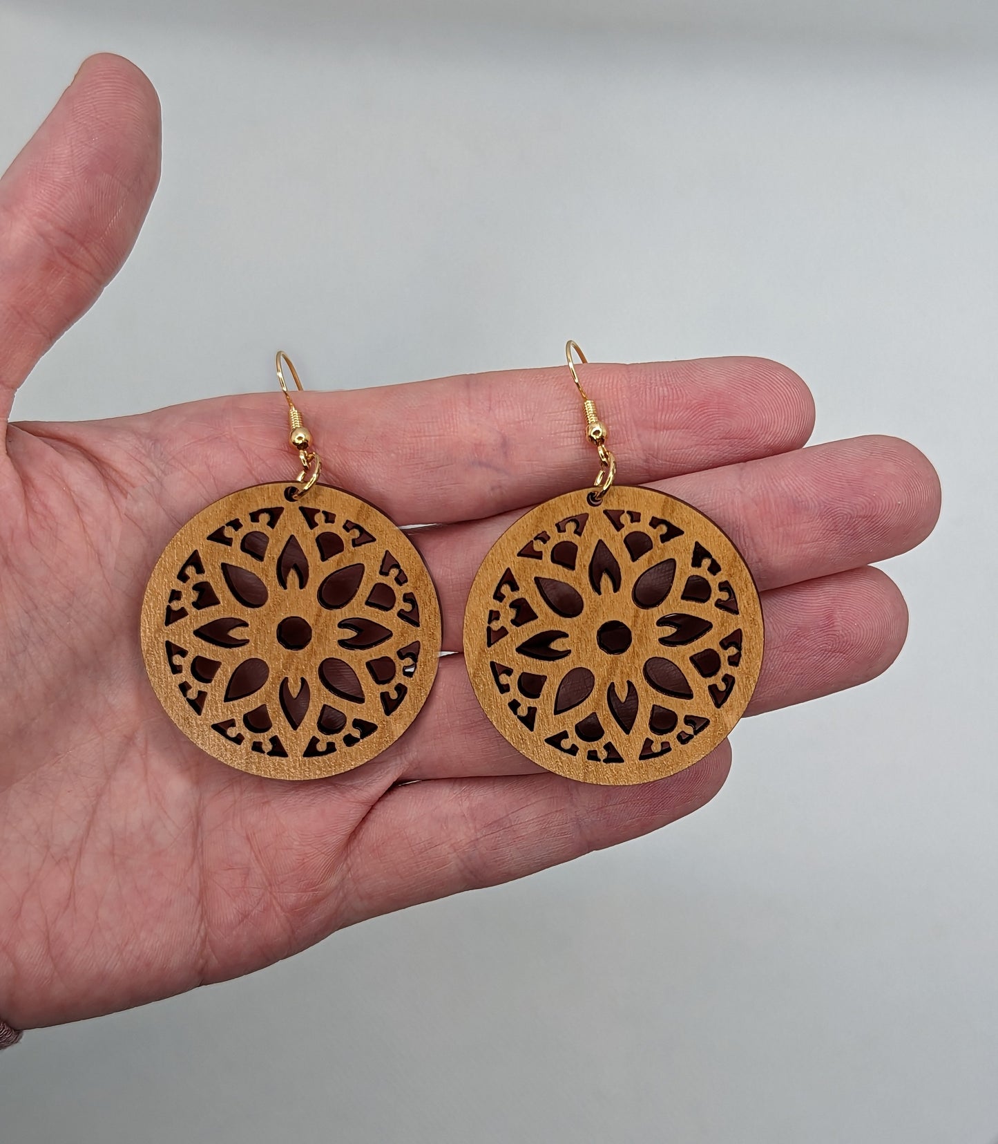Shows a pair of Floral Themed Circle earrings with an intricate pattern cut out of the middle, its on a hand showing the size against a white background, they are two finger widths in length and two wide. They are Cherry wood and have Gold fixings.