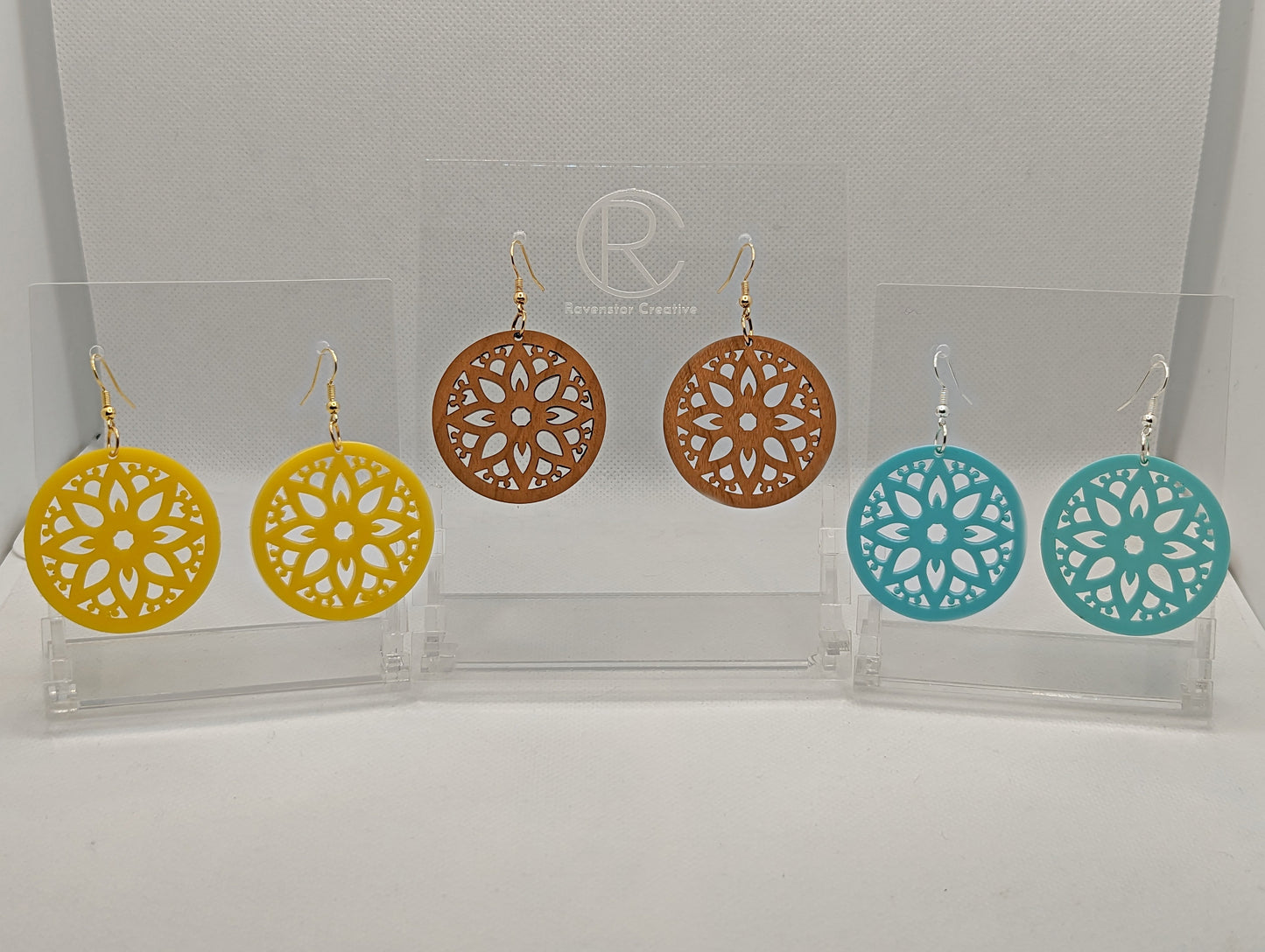 Three pairs of earrings that have ear wire hook fixings. They are Floral Themed Inspired Circle earrings with an intricate pattern. The colours are Cherry Wood, Yellow and Spearmint Acrylic. They are against a white background on see through stands.