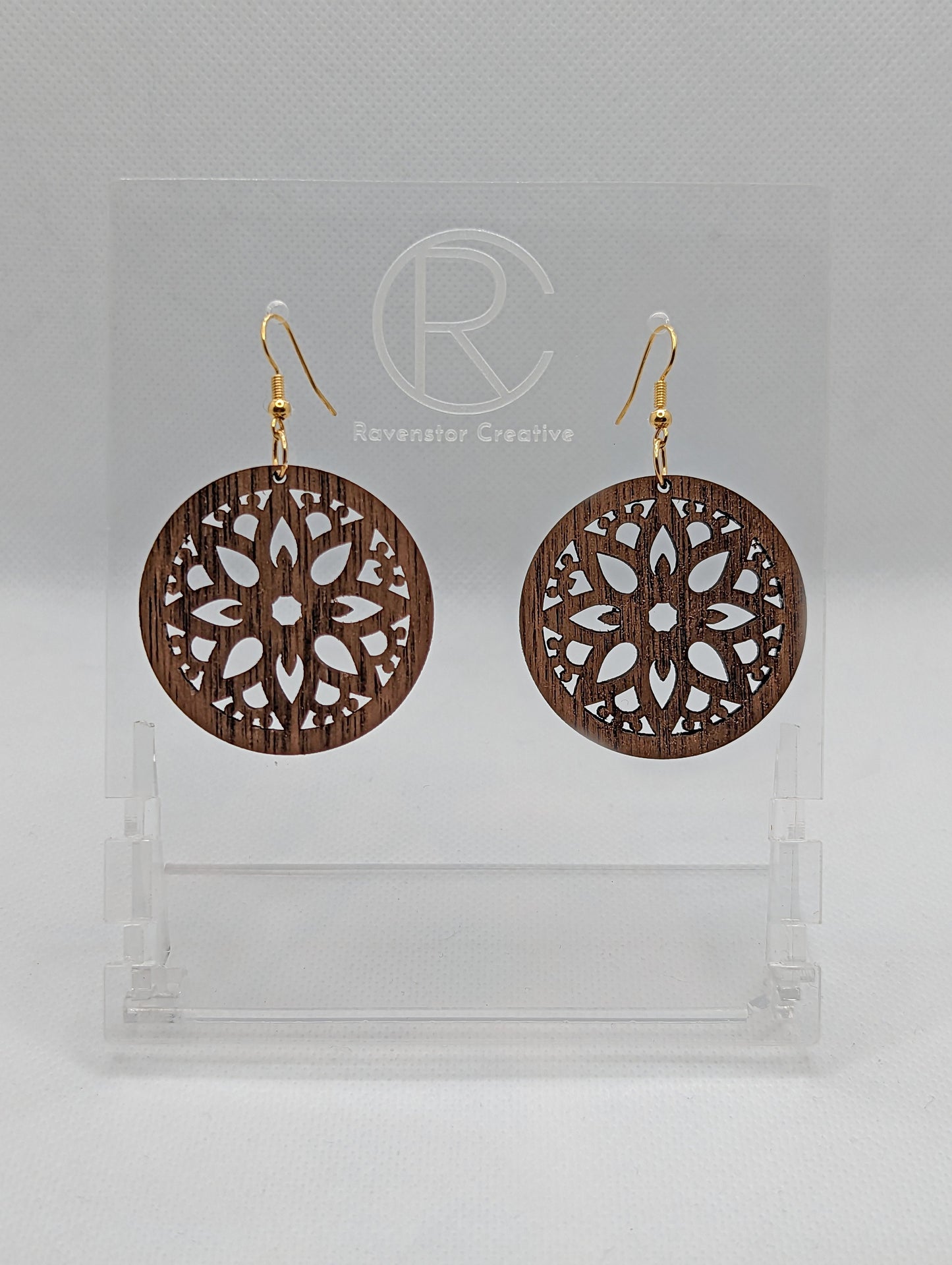 Shows a pair of earrings that have Gold ear wire hook fixings. They are Floral Themed Circle earrings with an intricate pattern cut out of the middle. They are walnut wood and are against a white background on see through stand.