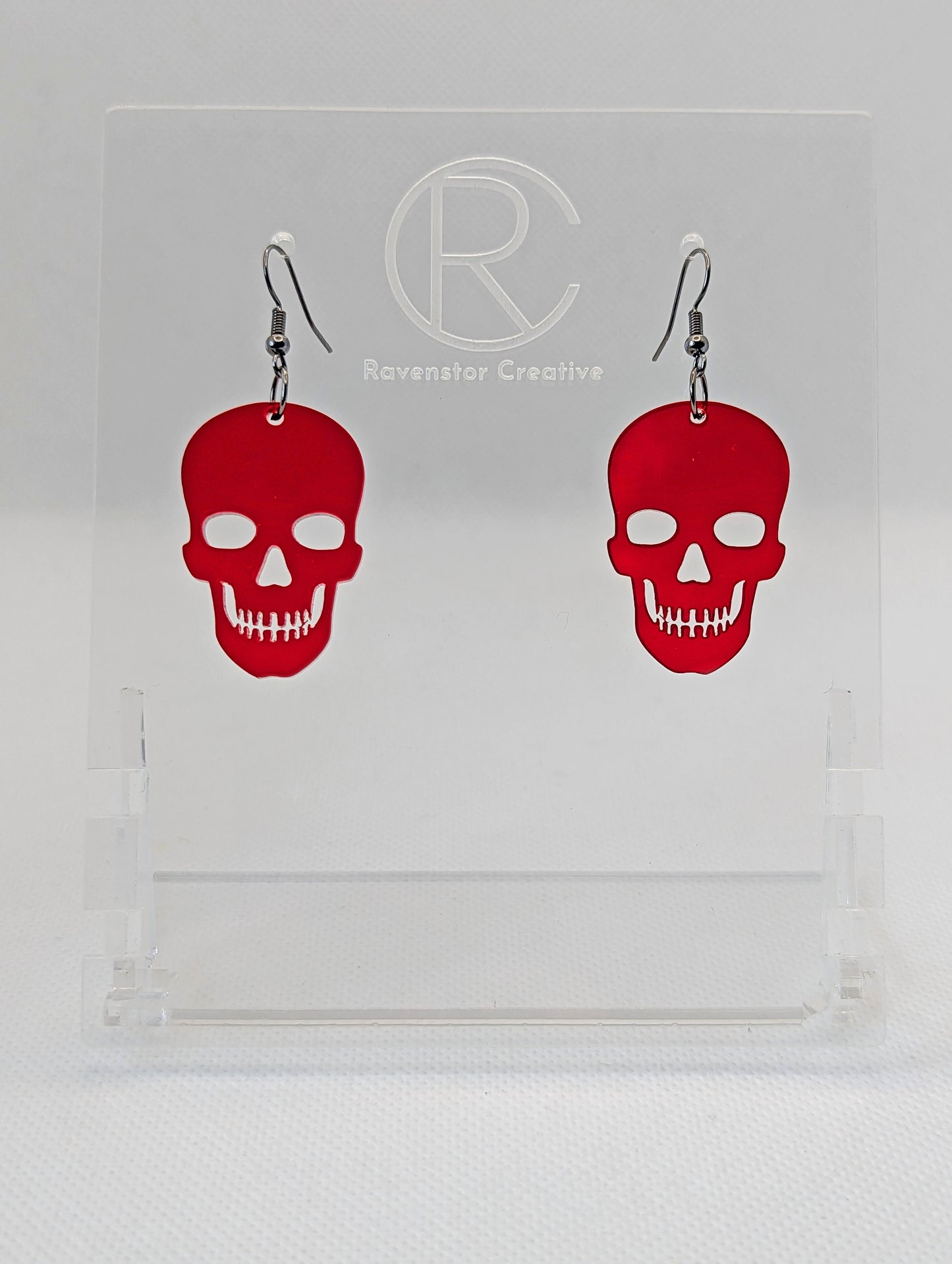 Shows a pair of earrings with black ear wire hook fixings. They are Scary Halloween Skull shapes. The colour of them is translucent red. They are on a see through stand that has the Ravenstor Creative Logo and its against a white background.