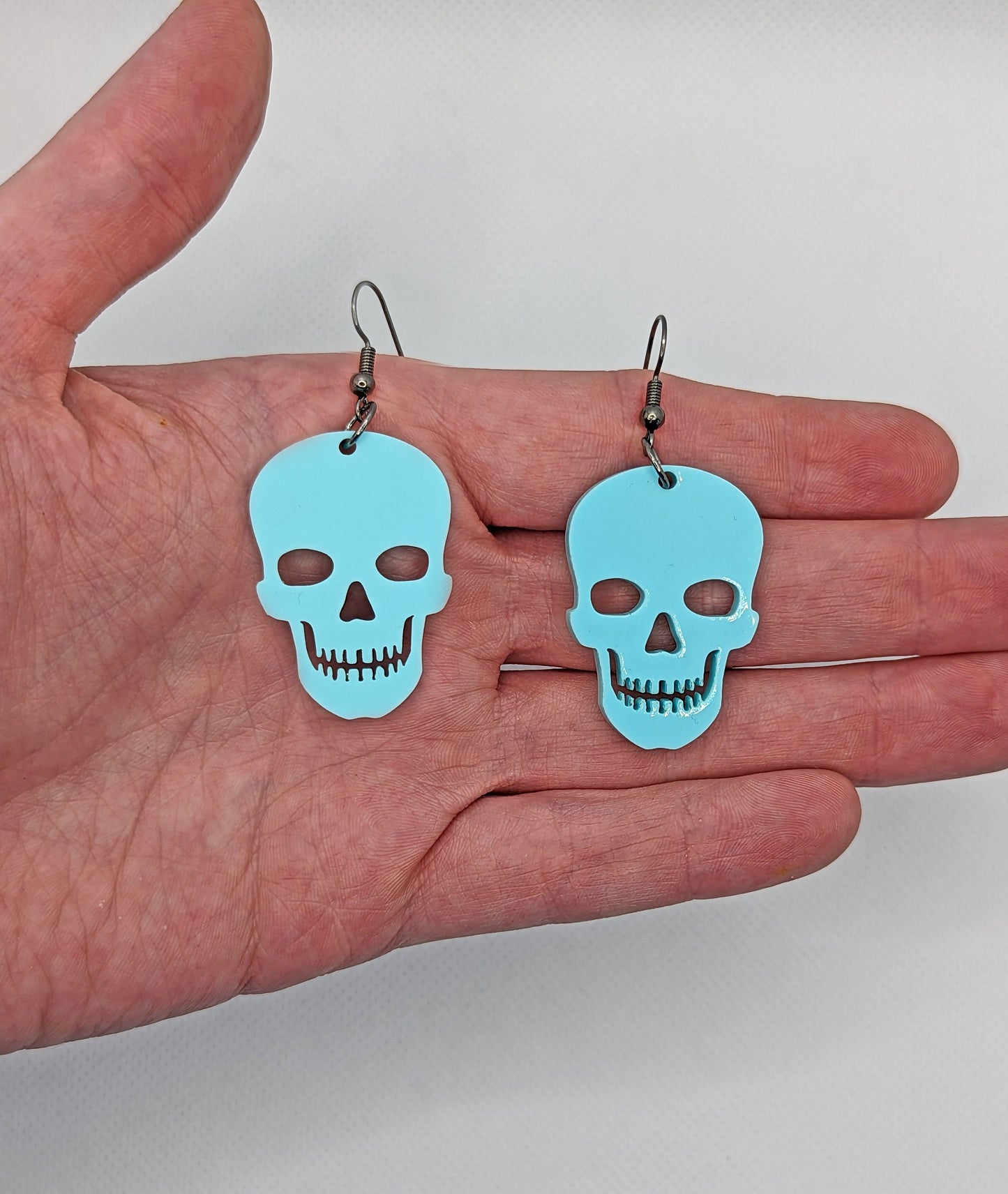 Image of a pair of Halloween Skull shaped earrings on a hand showing the size against a white background, they are two finger widths length and one and a half wide. They are spearmint and have black fixings.
