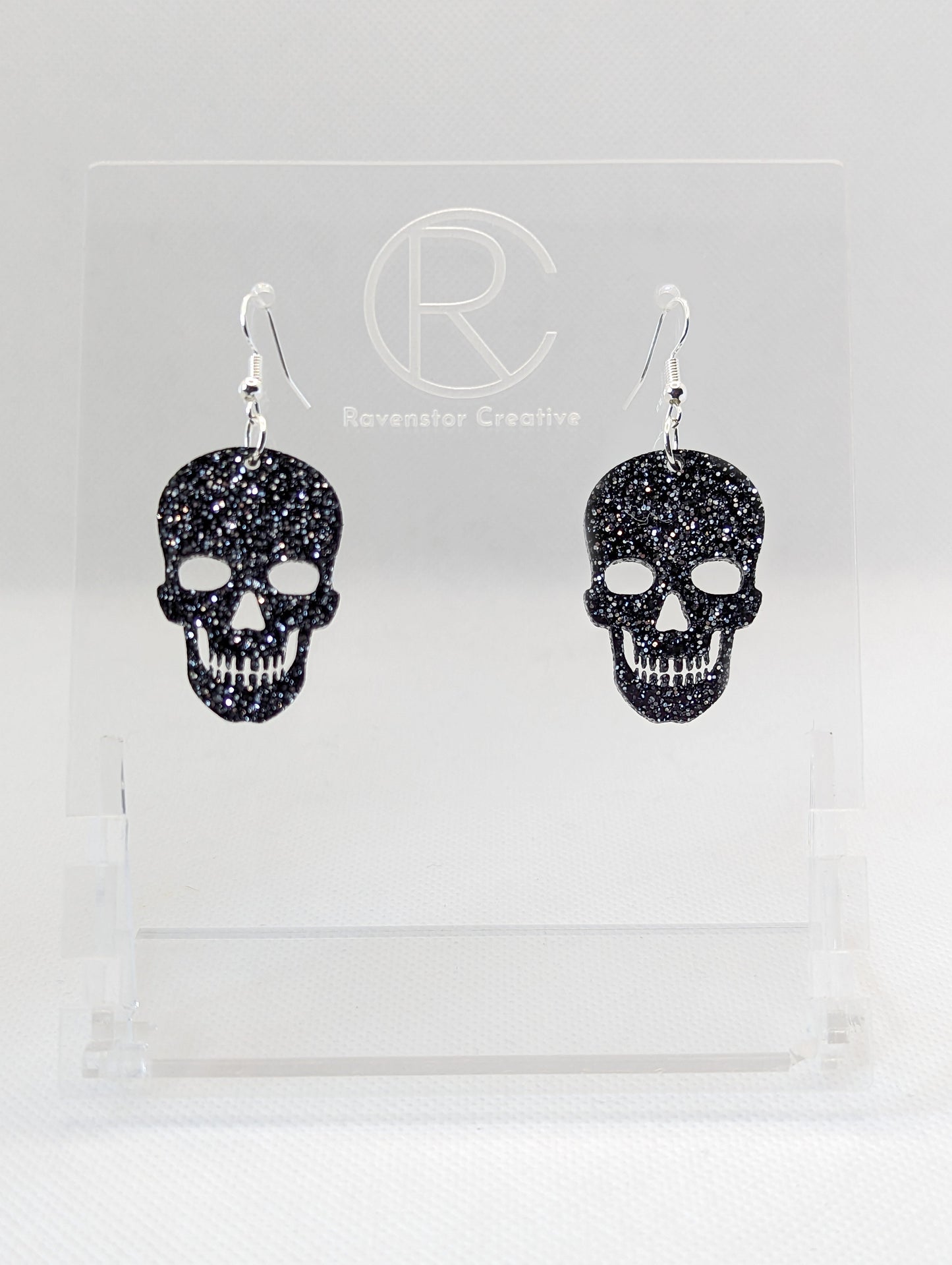 Shows a pair of earrings with Silver ear wire hook fixings. They are Scary Halloween Skull shapes. The colour of them is Black & Silver Glitter. They are on a see through stand that has the Ravenstor Creative Logo and its against a white background.