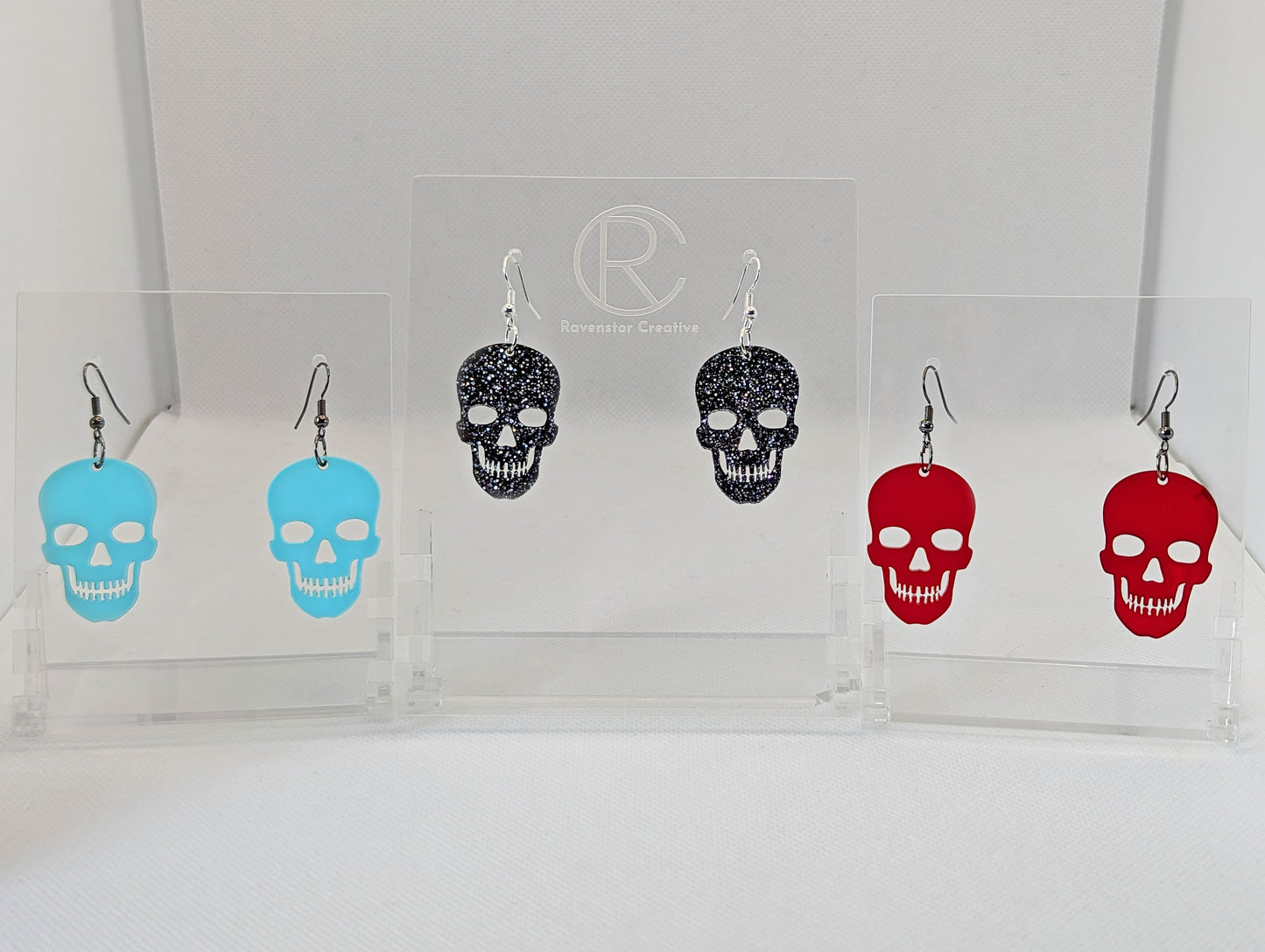 Shows 3 pairs of earrings with ear wire hook fixings. They are Scary Halloween skull shapes. The colours of them are spearmint, black & Silver glitter and translucent red. They are on see through stands against a white background