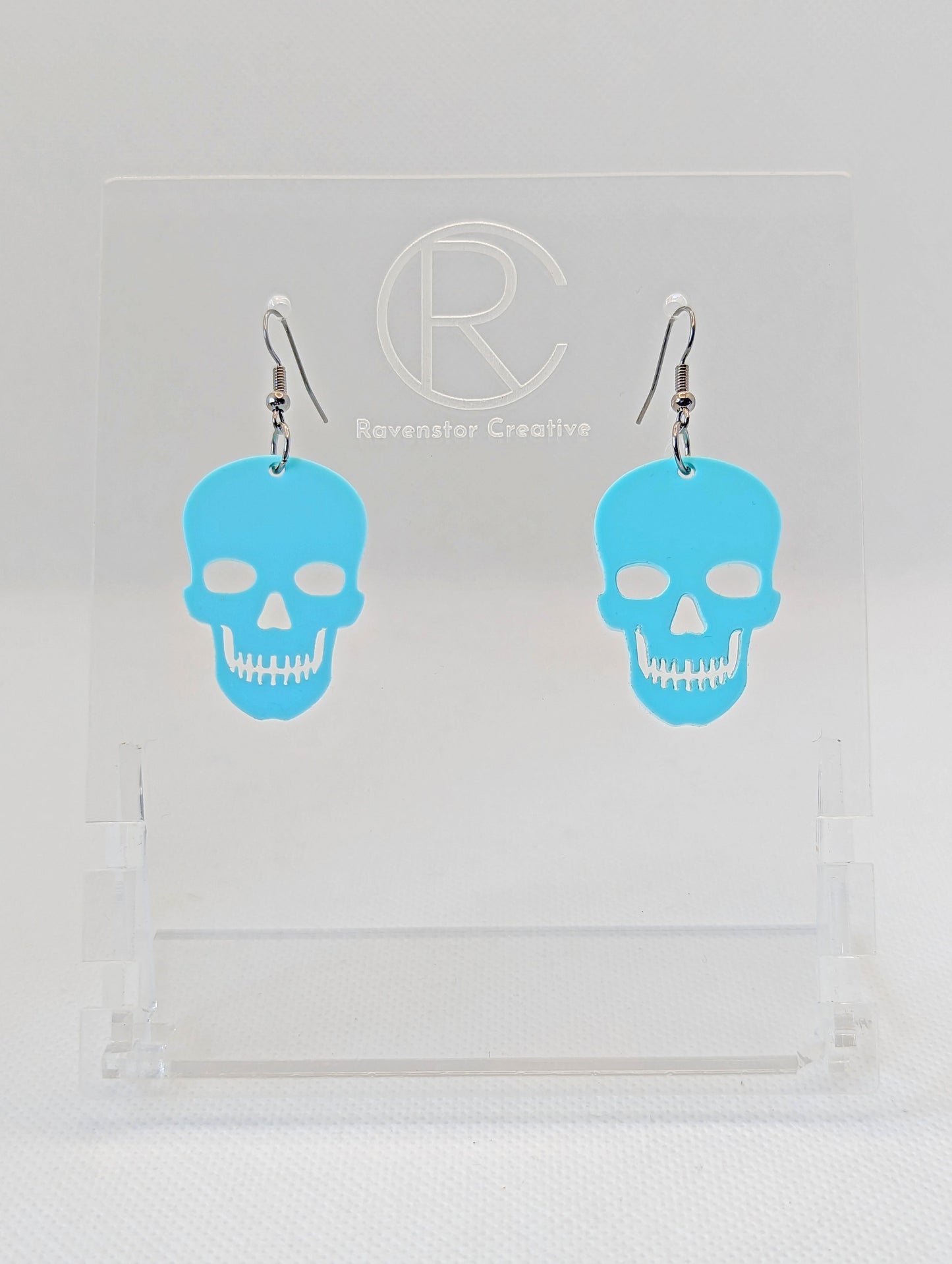 Shows a pair of earrings with black ear wire hook fixings. They are Scary Halloween Skull shapes. The colour of them is spearmint. They are on a see through stand that has the Ravenstor Creative Logo and its against a white background.