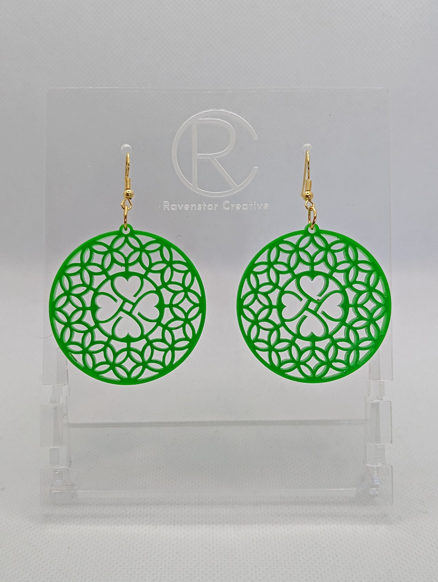 Shows a pair of acrylic earrings that have gold ear wire hook fixings. They are circles with a shamrock four leaf clover design. They are Green in colour and are against a white background on see through stand with the Ravenstor Creative logo on it.
