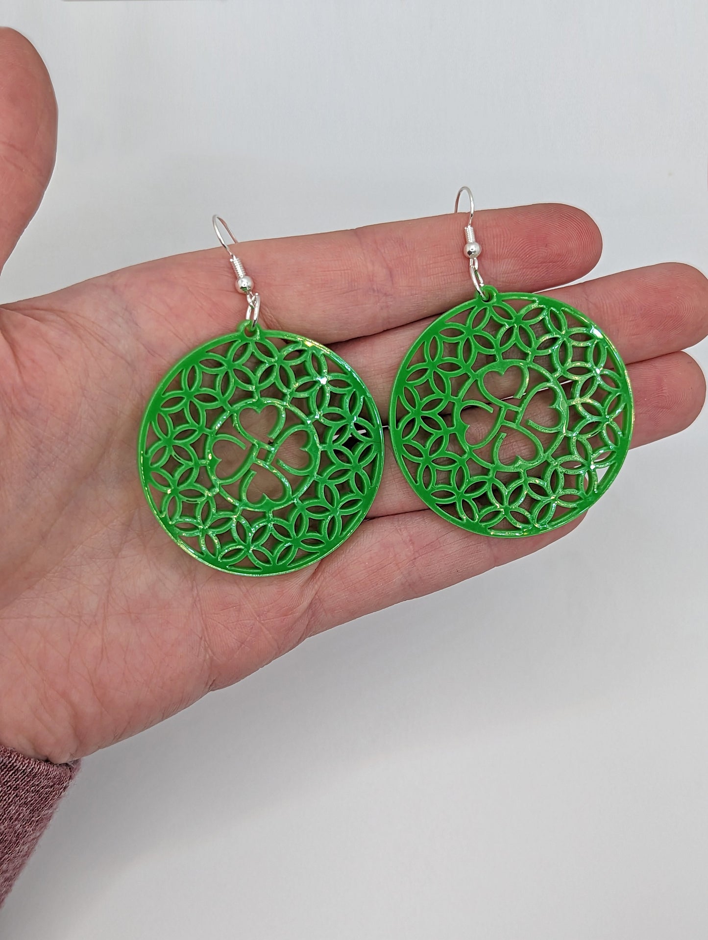 Shows a pair of acrylic earrings that have silver ear wire hook fixings. They are circles with a shamrock four leaf clover design. They are against a hand showing the size of 3 finger widths in length and same again wide.