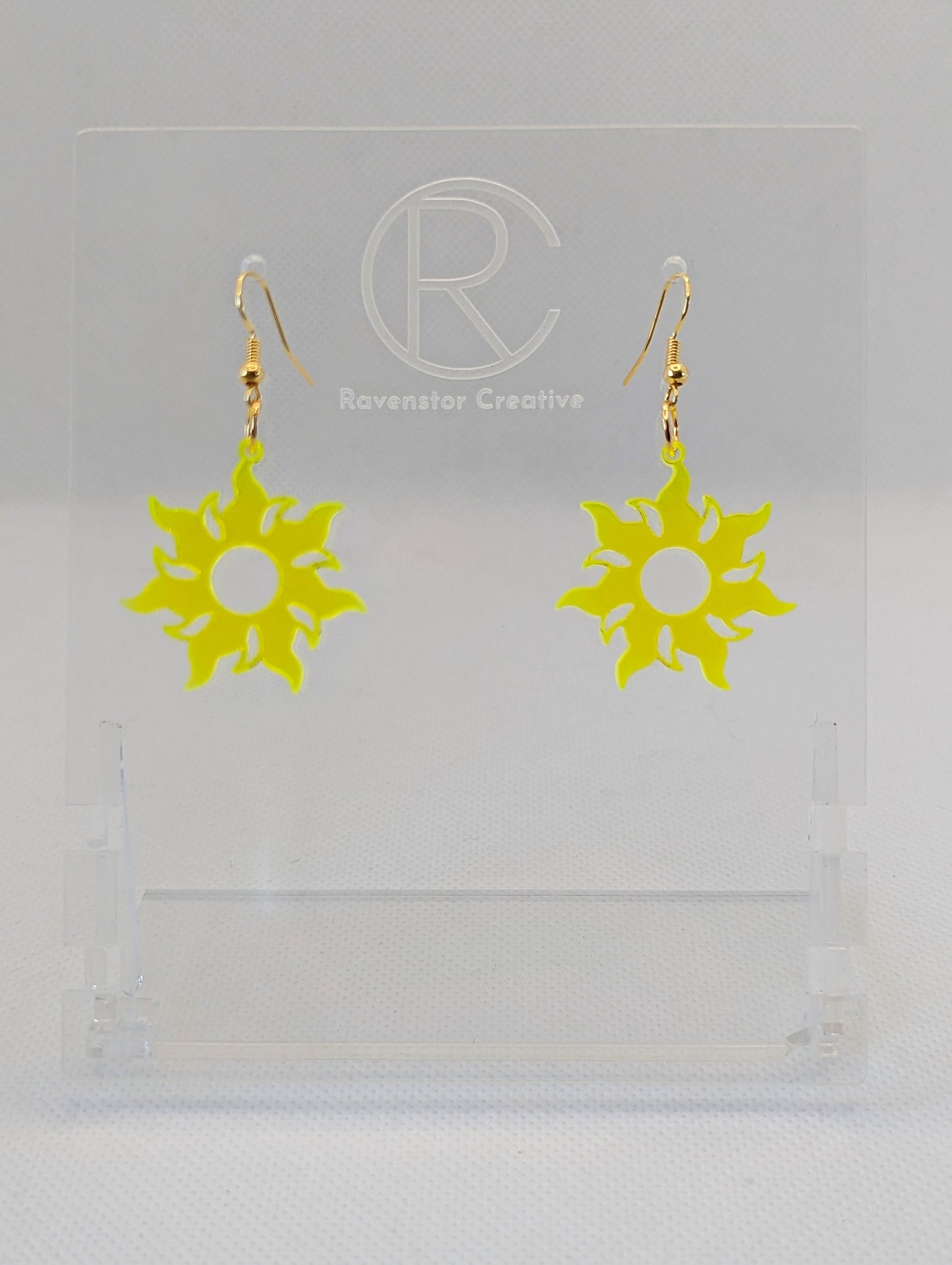 Shows a pair of earrings that have gold ear wire hook fixings. They are shining tangled sun shapes. They are neon yellow in colour and are against a white background on see through stand with the Ravenstor Creative logo on it.
