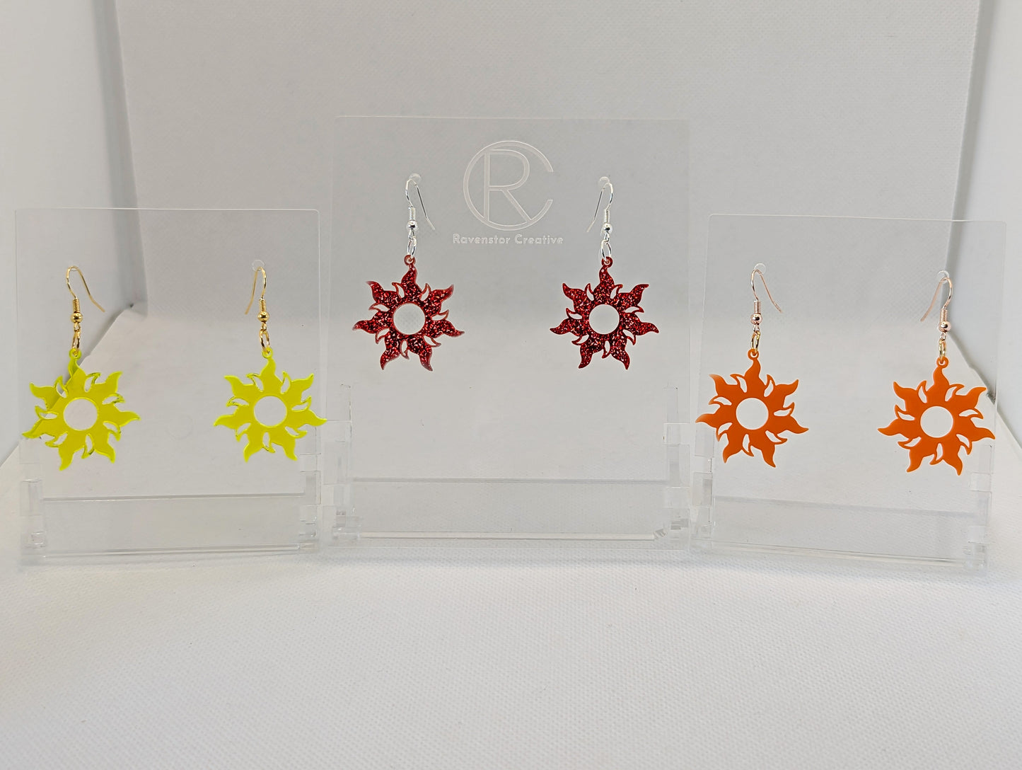 Shows three pairs of earrings that have ear wire hook fixings. They are shining tangled sun shaped. The colours are neon yellow, red glitter and orange. They are against a white background on see through stand with the ravenstor creative logo on.