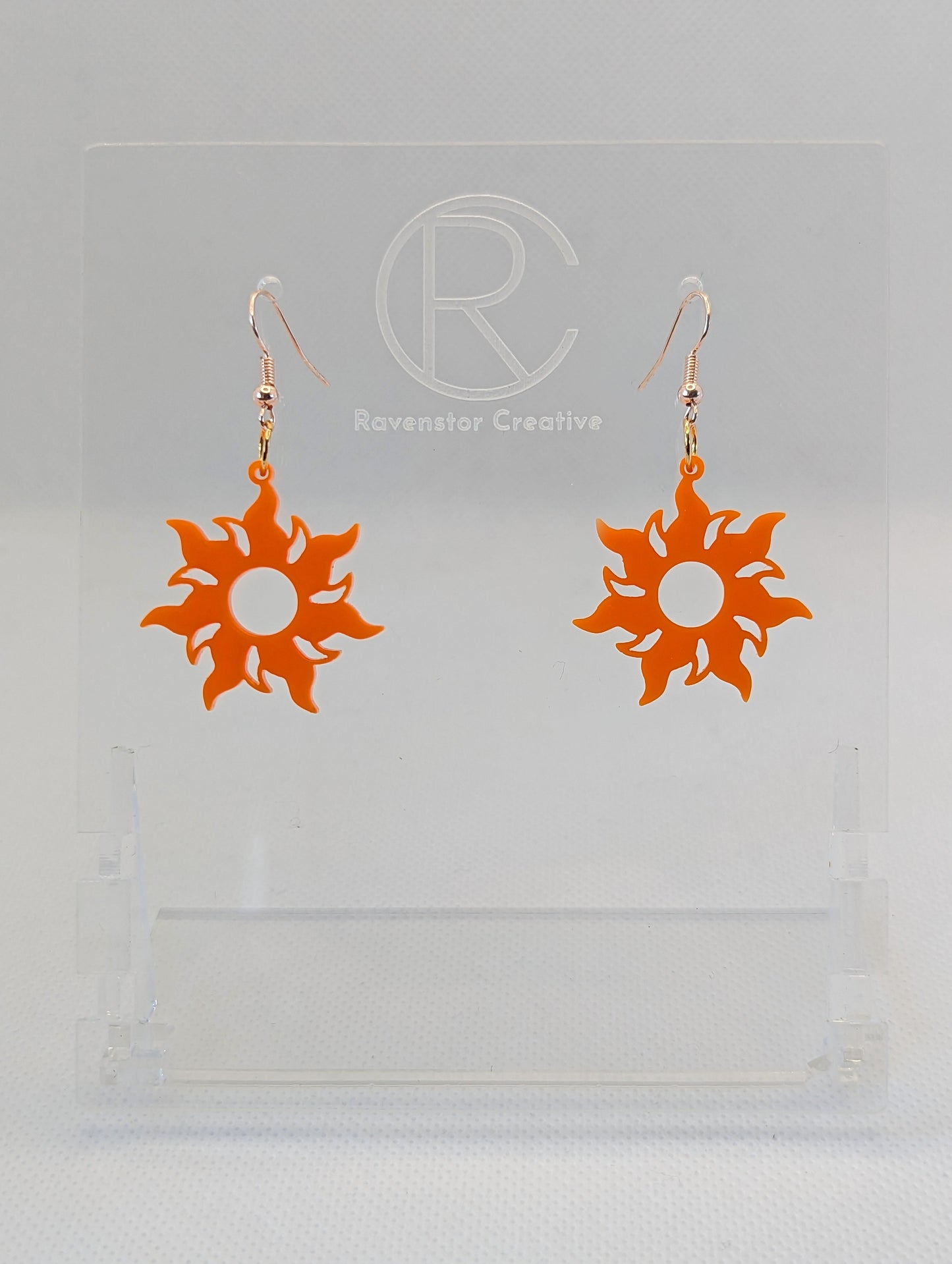 Shows a pair of earrings that have rose gold ear wire hook fixings. They are shining tangled sun shapes. They are orange in colour and are against a white background on see through stand with the Ravenstor Creative logo on it.