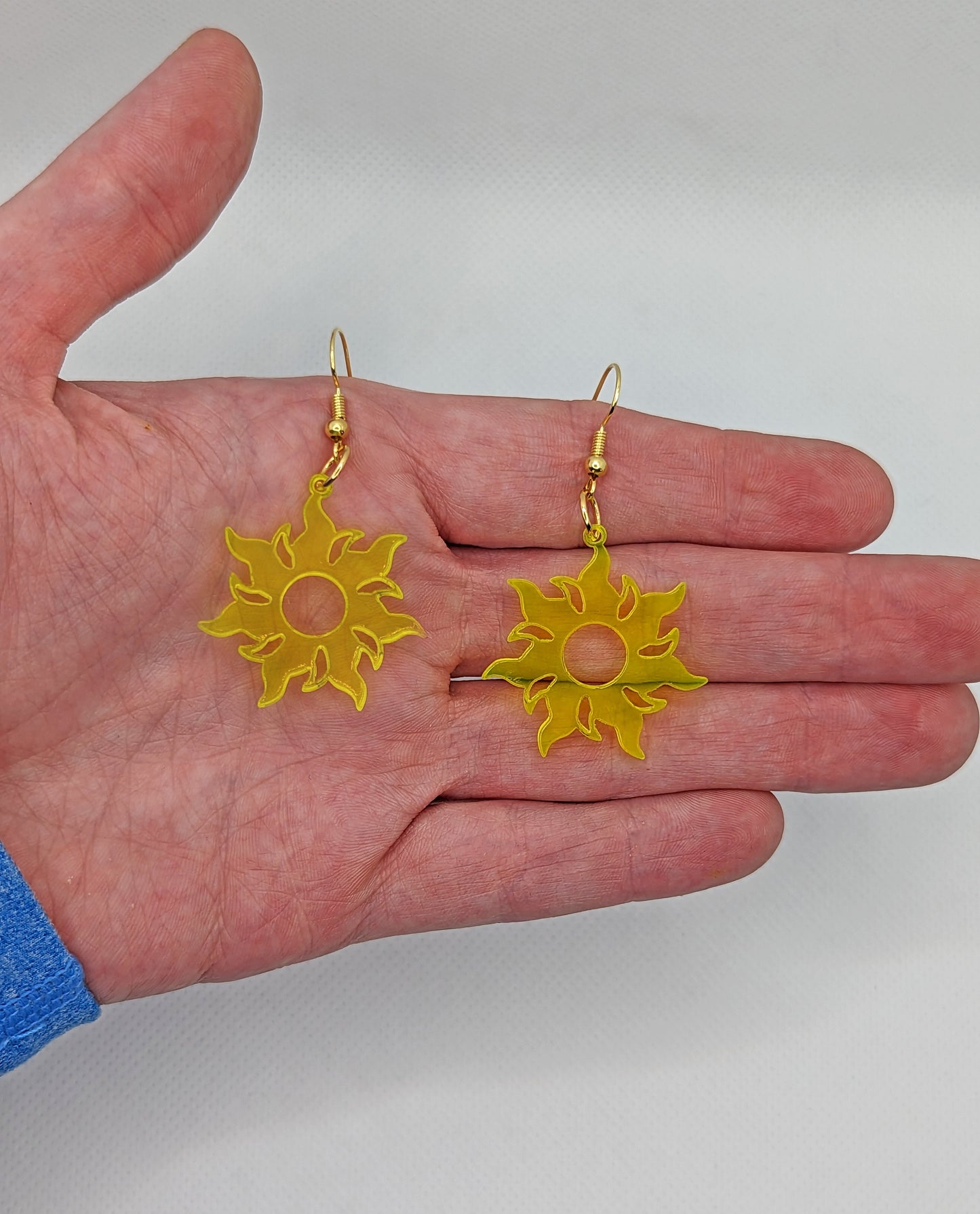 Image of a pair of Shining Tangled Sun shaped earrings on a hand showing the size against a white background, they are one and a half finger widths length and same again wide. They are neon yellow and have gold fixings.
