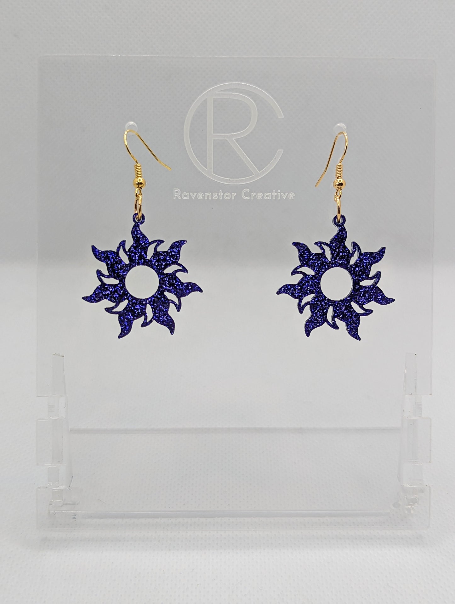 Shows a pair of earrings that have gold ear wire hook fixings. They are shining tangled sun shapes. They are purple glitter in colour and are against a white background on see through stand with the Ravenstor Creative logo on it.