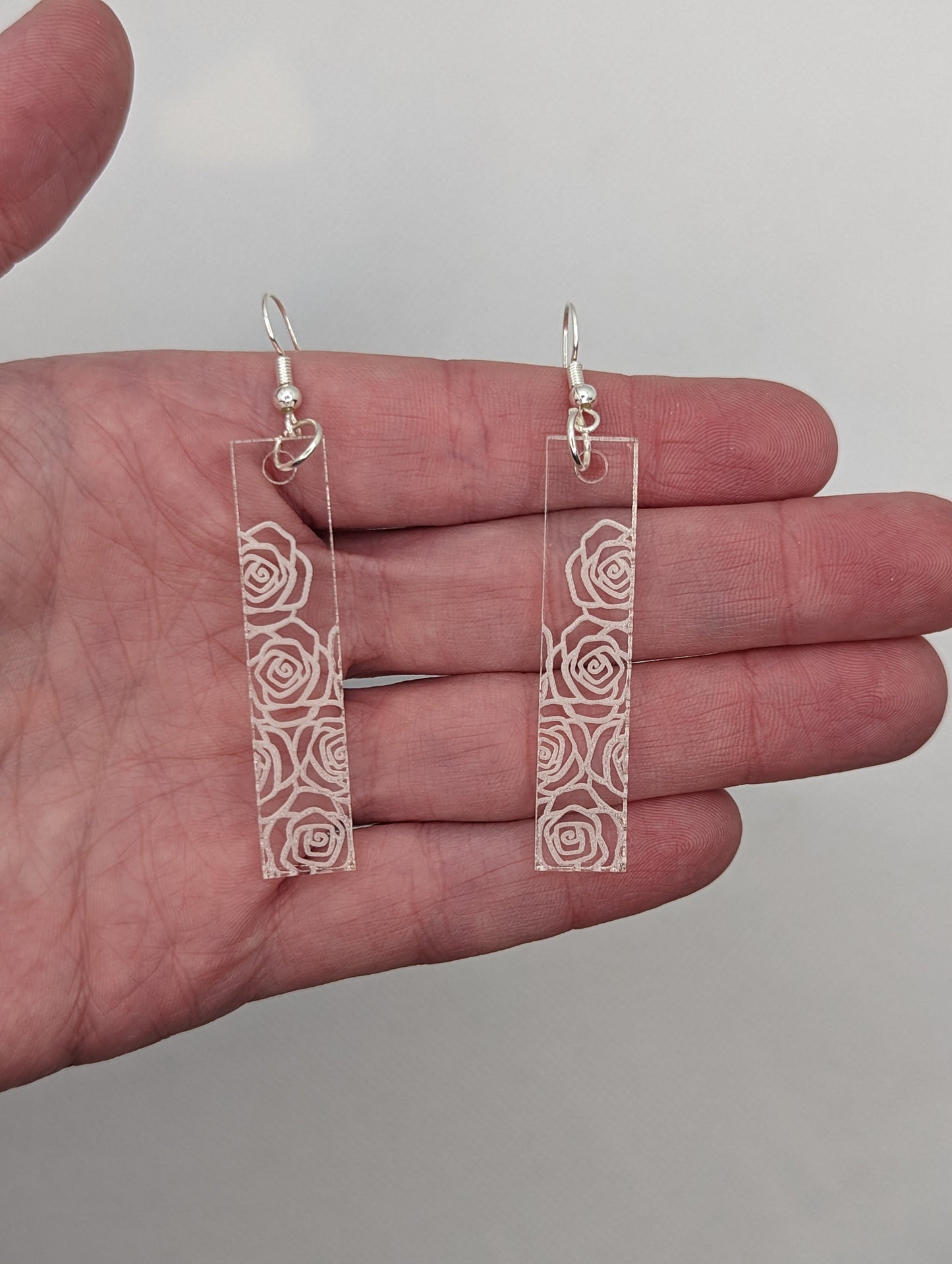 Image of a pair of long slim rectangle earrings with silver fixings on a hand showing the size, they are three finger widths in length and one wide. They are clear/see through and are engraved with roses.