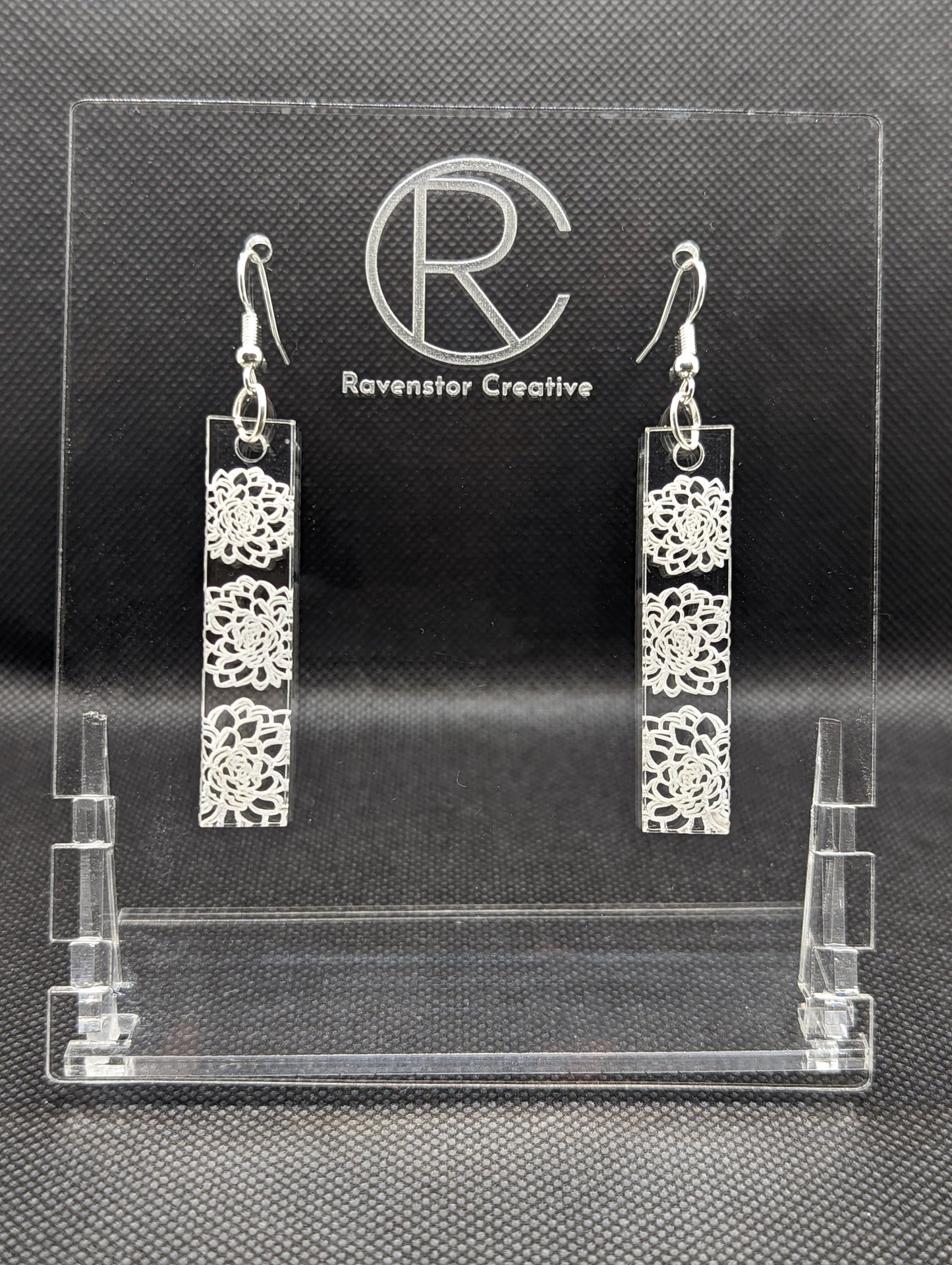 Shows a pair of long slim Floral themed rectangle acrylic clear/see through earrings that have silver ear wire hook fixings. Each is engraved with three Dahlias / Flowers . They are against a dark background on see through stand.