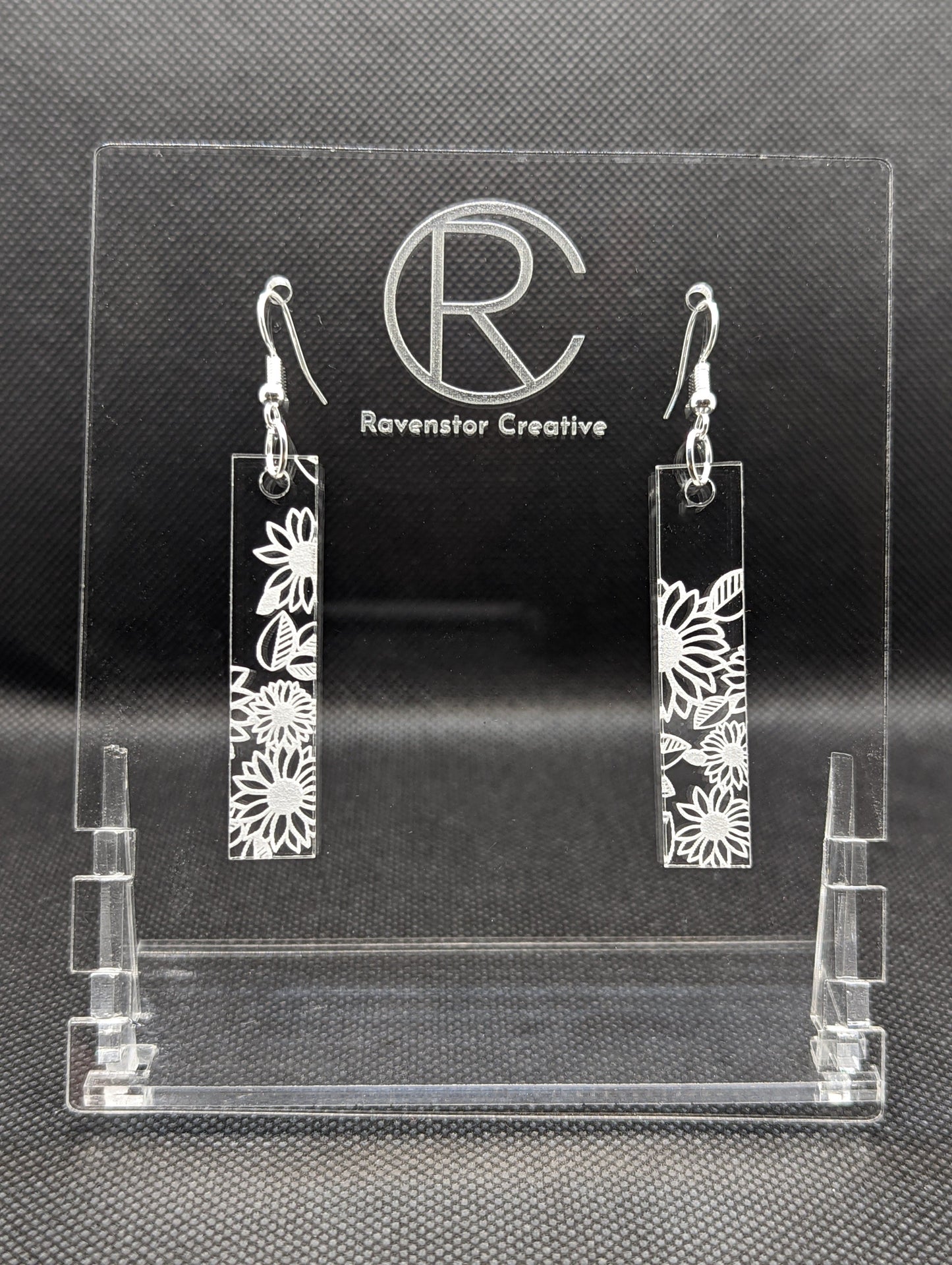 Shows a pair of long slim Floral themed rectangle acrylic clear/see through earrings that have silver ear wire hook fixings. Each is engraved with Daises / Flowers . They are against a dark background on see through stand.