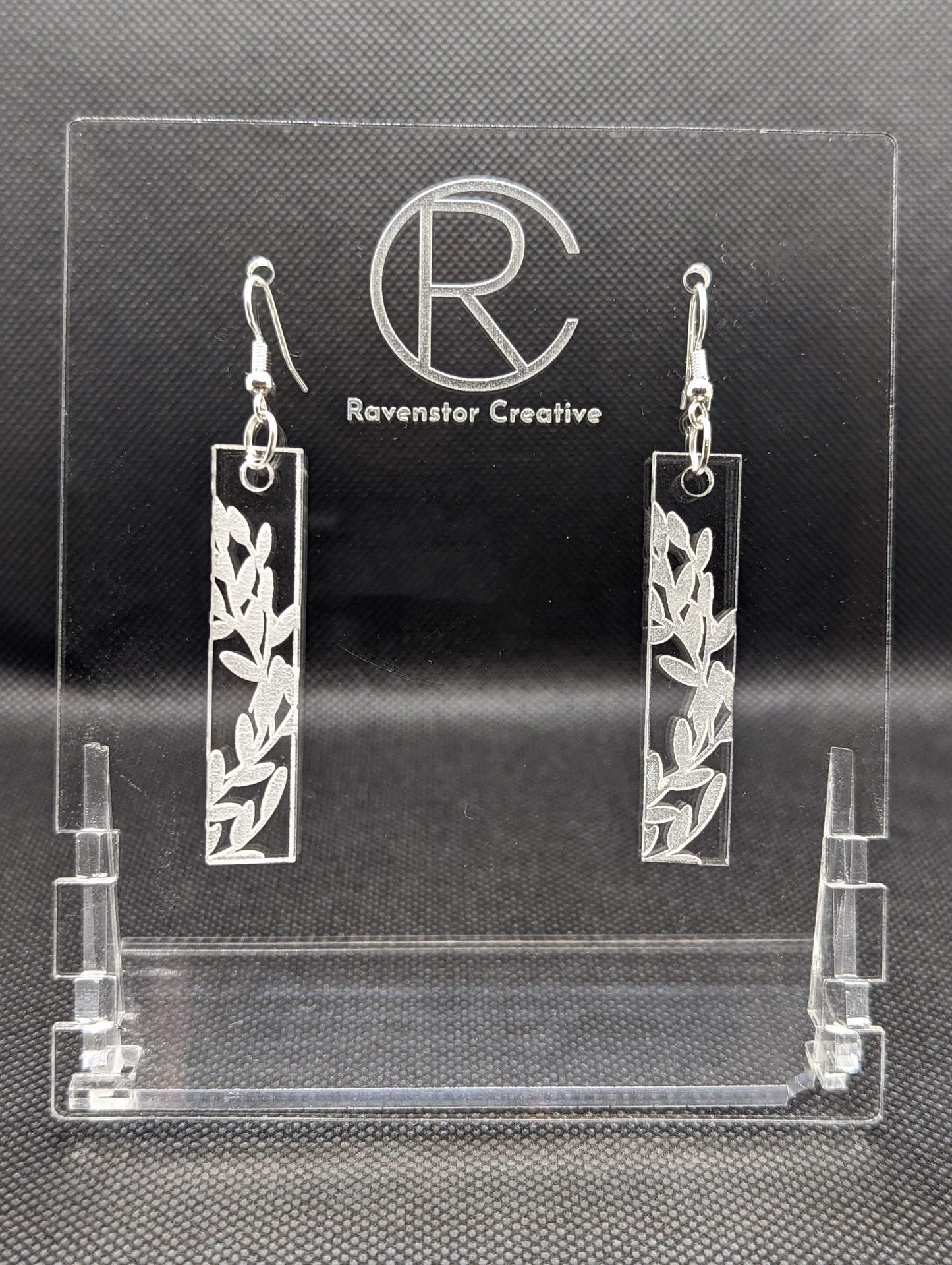 Shows a pair of long slim Floral themed rectangle acrylic clear/see through earrings that have silver ear wire hook fixings. Each is engraved with Bay Leaves / Flowers . They are against a dark background on see through stand.