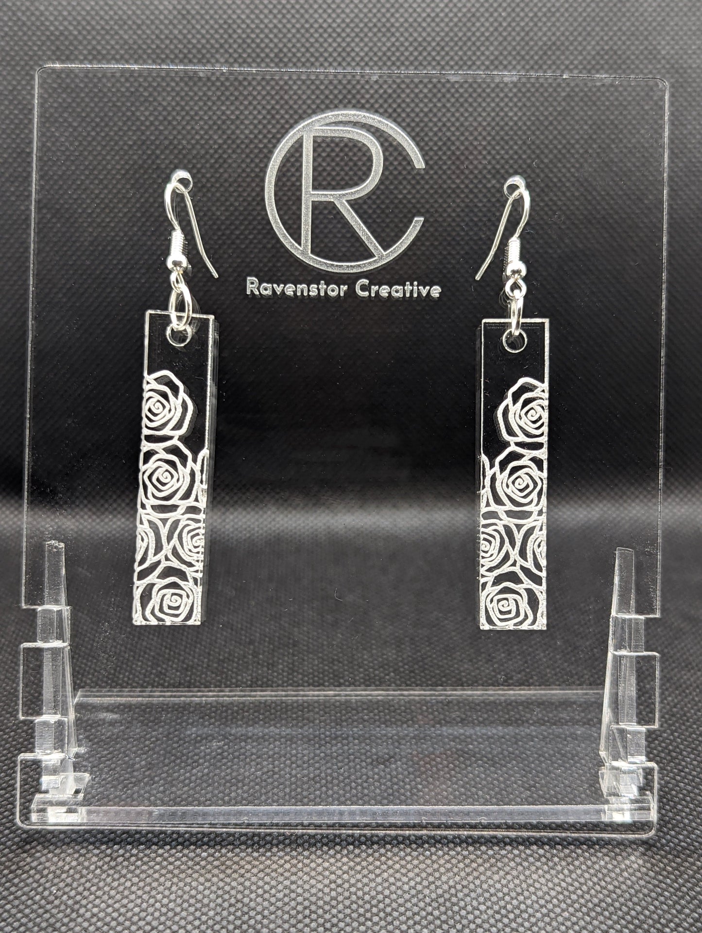 Shows a pair of long slim Floral themed rectangle acrylic clear/see through earrings that have silver ear wire hook fixings. Each is engraved with Roses / Flowers . They are against a dark background on see through stand.