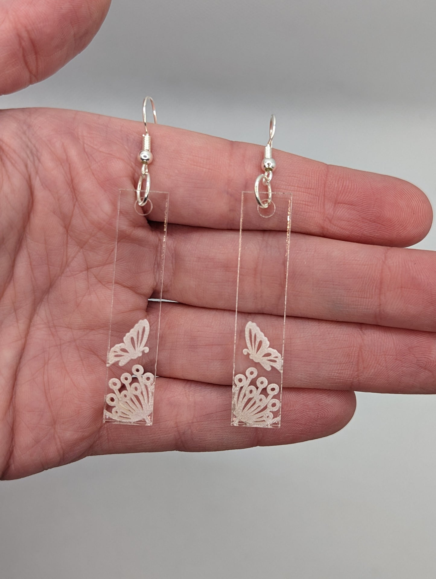 Image of a pair of long slim rectangle earrings with silver fixings on a hand showing the size, they are three finger widths in length and one wide. They are clear/see through and are engraved with a butterfly landing on a flower.