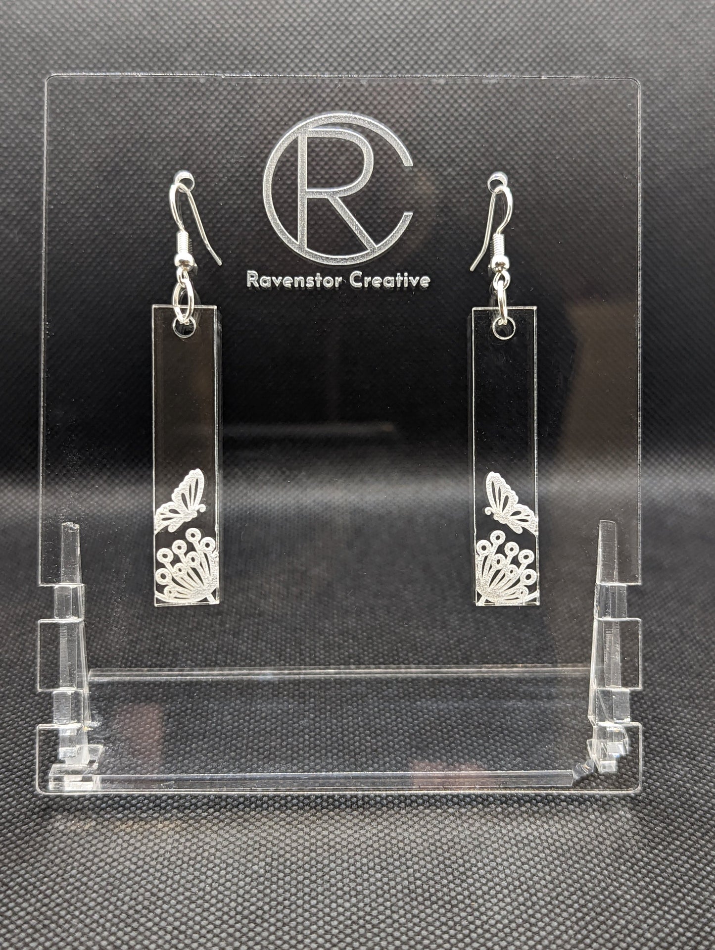 Shows a pair of long slim rectangle acrylic clear/see through earrings that have silver ear wire hook fixings. Each is engraved with a butterfly landing on a flower. They are against a dark background on see through stand.