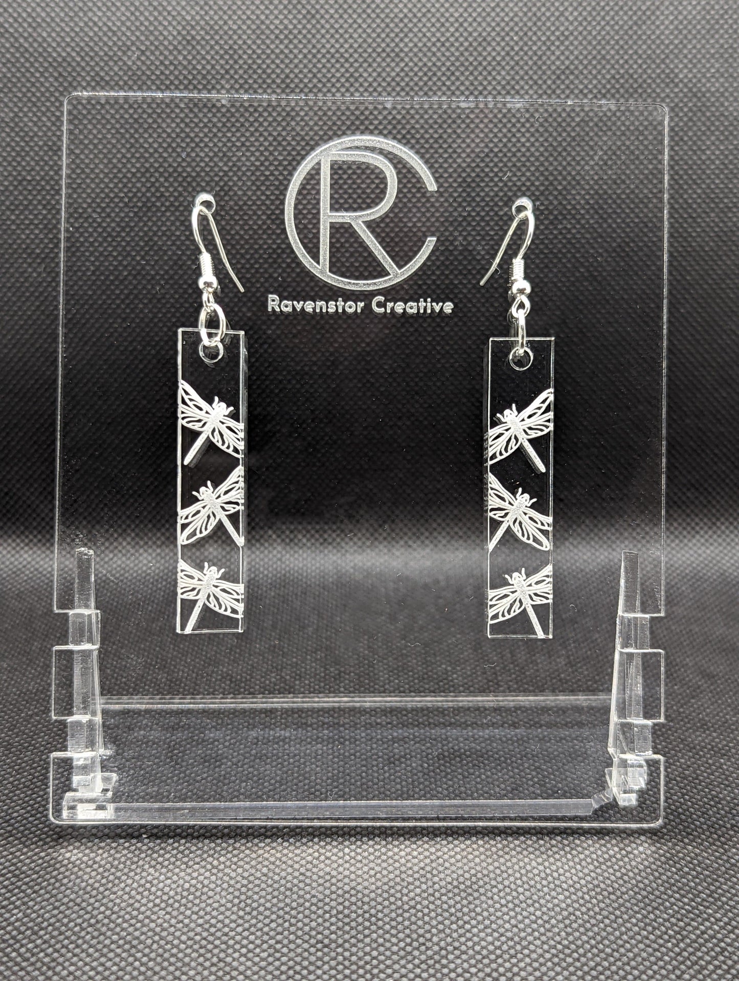 Shows a pair of long slim rectangle acrylic clear/see through earrings that have silver ear wire hook fixings. Each is engraved with three dragon flies. Dragon fly themed and are against a dark background on see through stand.