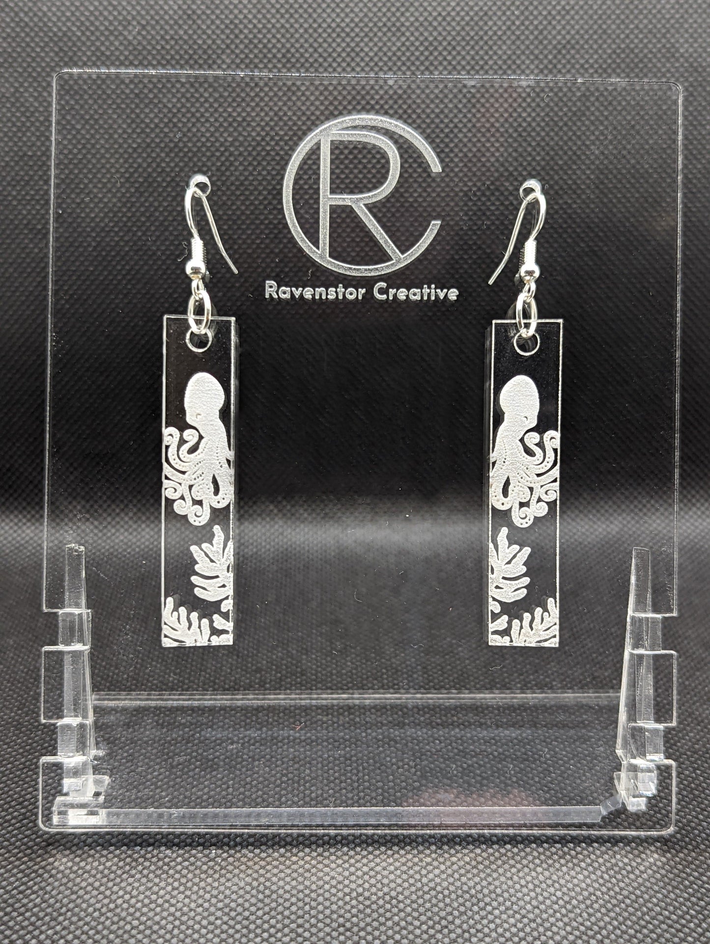 Shows a pair of long slim rectangle acrylic clear/see through earrings that have silver ear wire hook fixings. Each is engraved with an Octopus above some seaweed. They are against a dark background on see through stand.