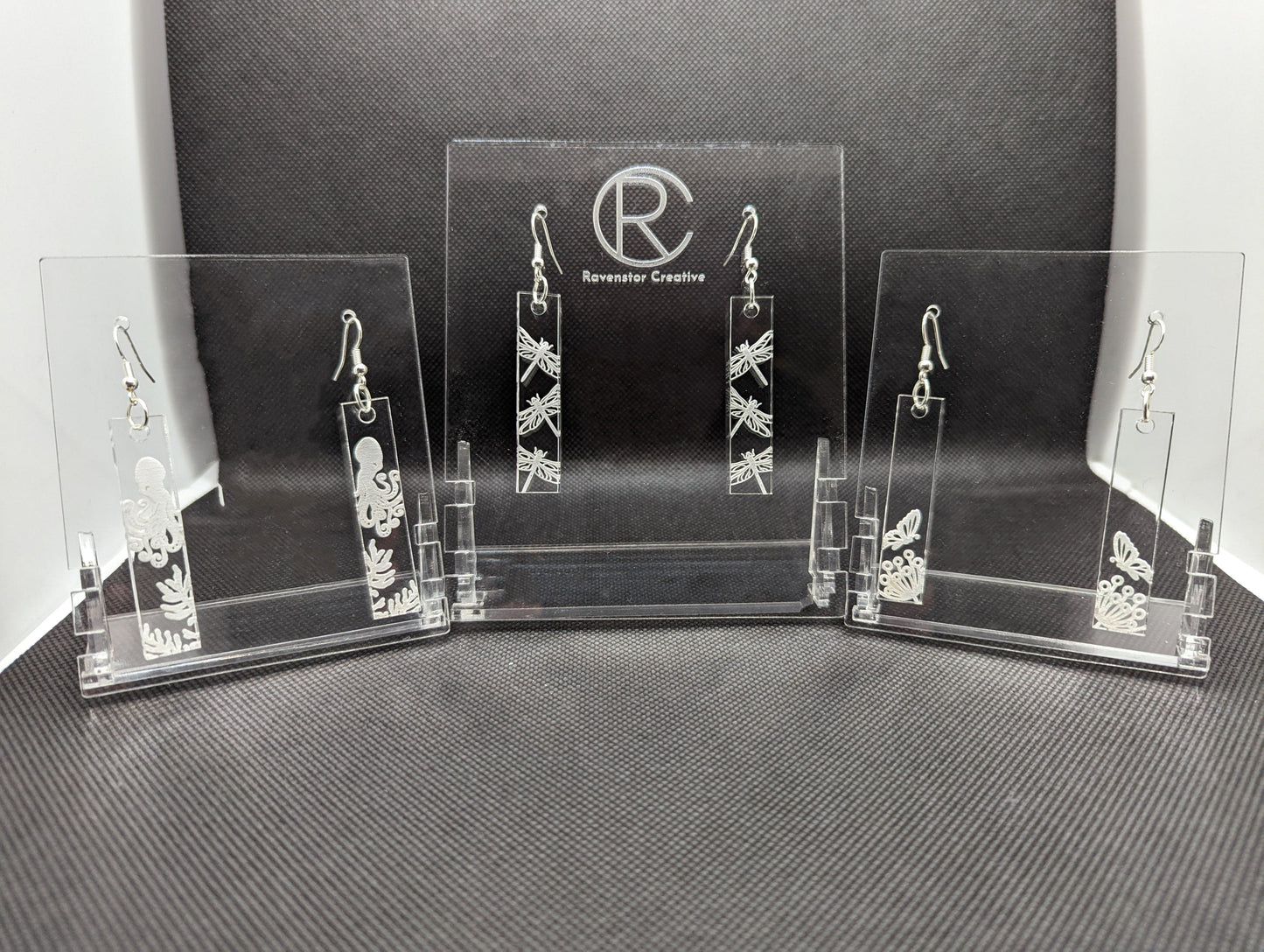 Shows three pairs of slim rectangle see through clear earrings with silver fixings that are engraved with nature scenes. One is an octopus with seaweed under it, One pair has three dragon flies on each, one pair has butterflies landing on a flower.