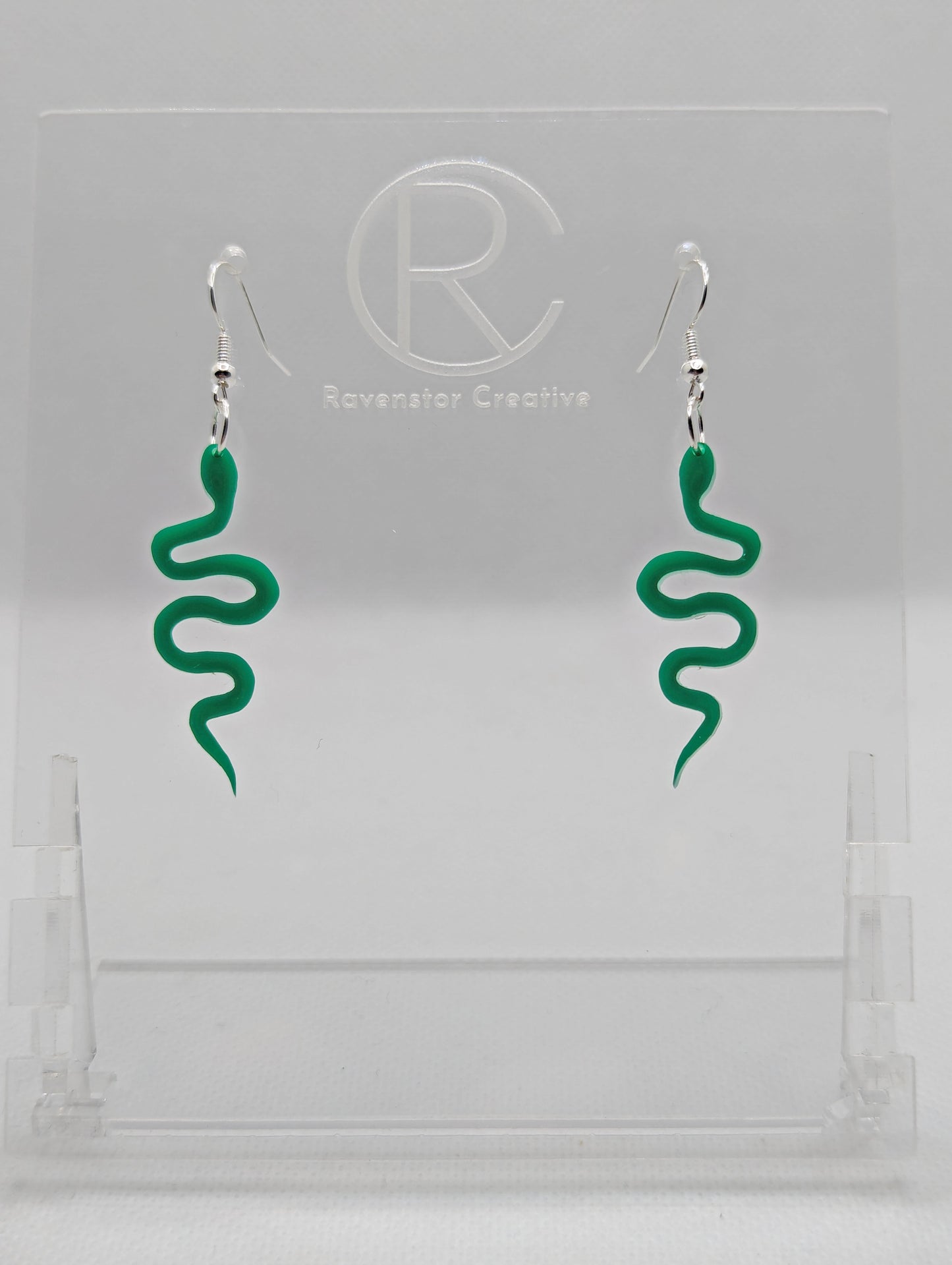 Shows a pair of acrylic earrings with silver ear wire hook fixings. They are snake shapes. The colour is Green. They are against a white background on a clear stand with the Ravenstor Creative logo on it.