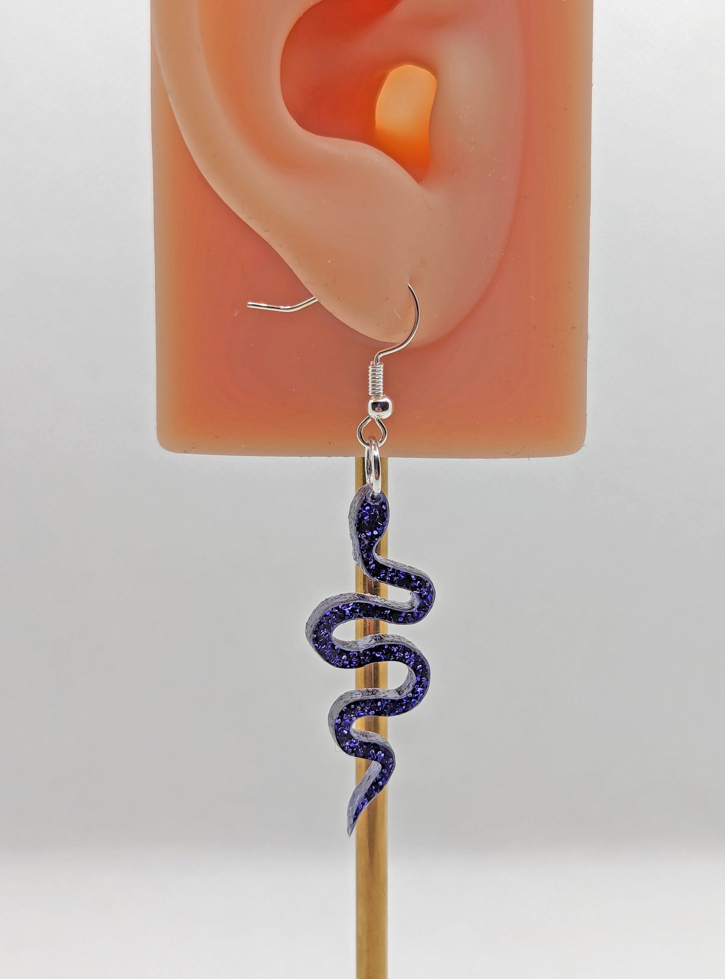 Shows a pair of Acrylic earrings with gold ear wire hook fixings. They are snake shapes. The colour is Purple Glitter. They are against an ear to show the size.