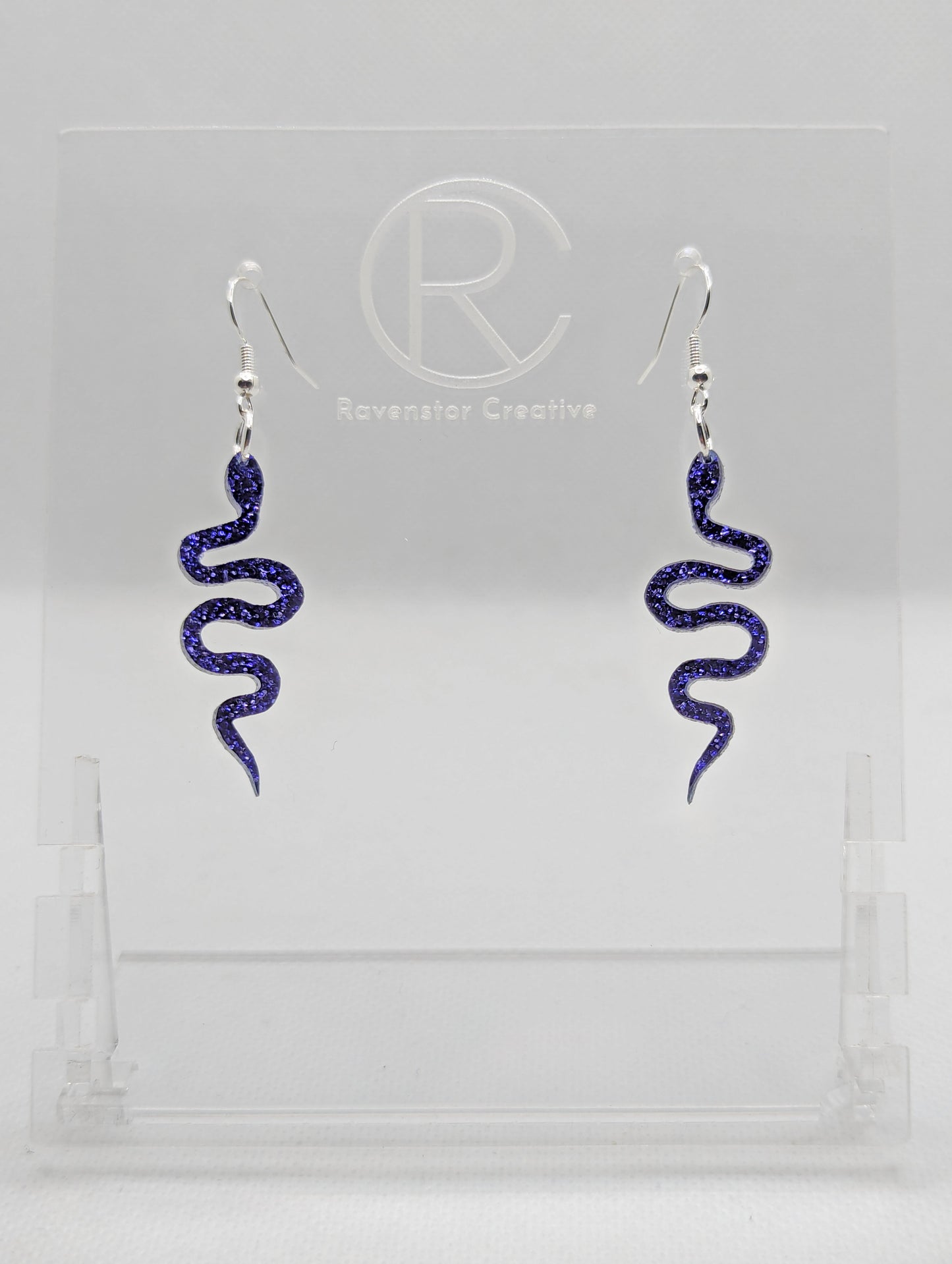 Shows a pair of acrylic earrings with silver ear wire hook fixings. They are snake shapes. The colour is Purple Glitter. They are against a white background on a clear stand with the Ravenstor Creative logo on it.