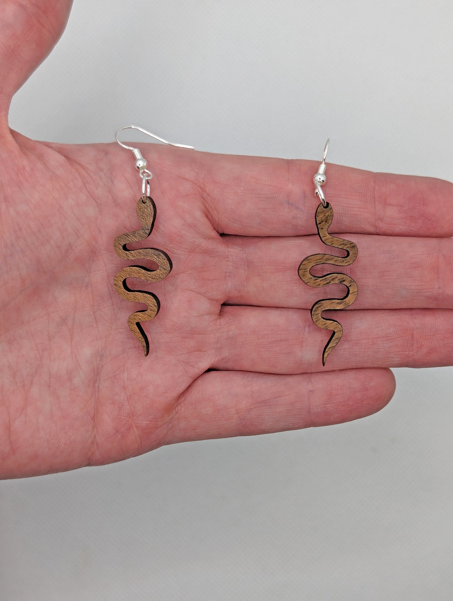 Shows a pair of wooden earrings with gold ear wire hook fixings. They are snake shapes. The colour is Walnut Wood. They are against a hand showing the size of two and a half finger widths in length and one wide.