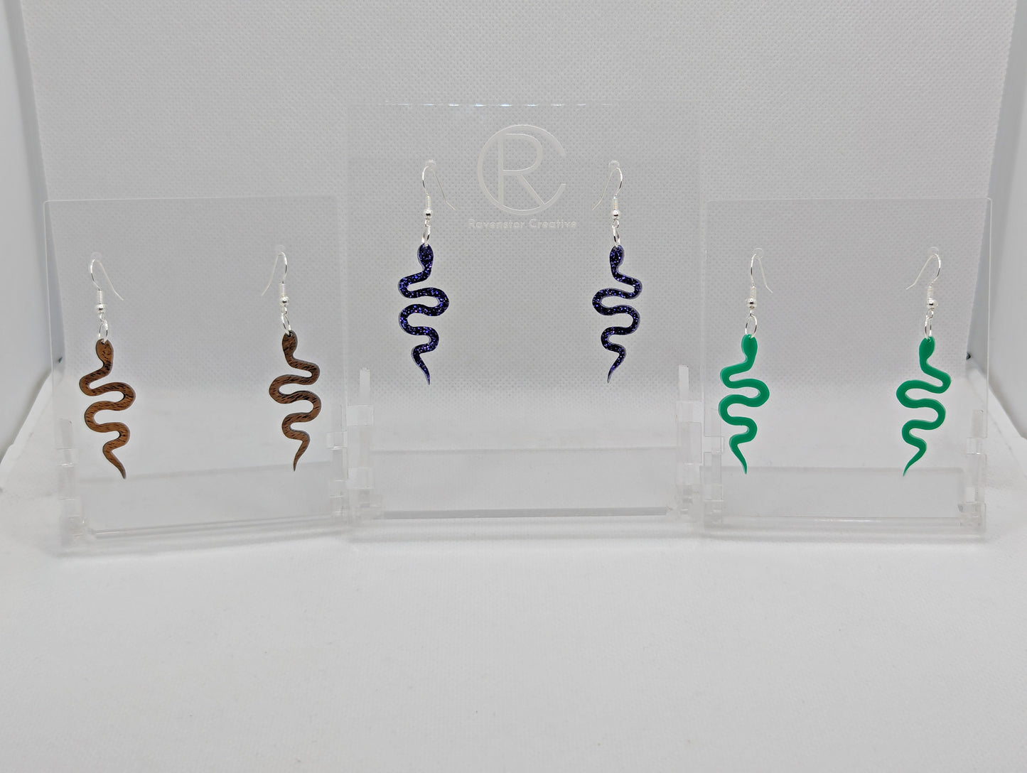 Shows three pairs of earrings with ear wire hook fixings. They are snake shapes. The colours are Walnut Wood, Purple Glitter Acrylic and Green Acrylic. They are against a white background on a clear stand.