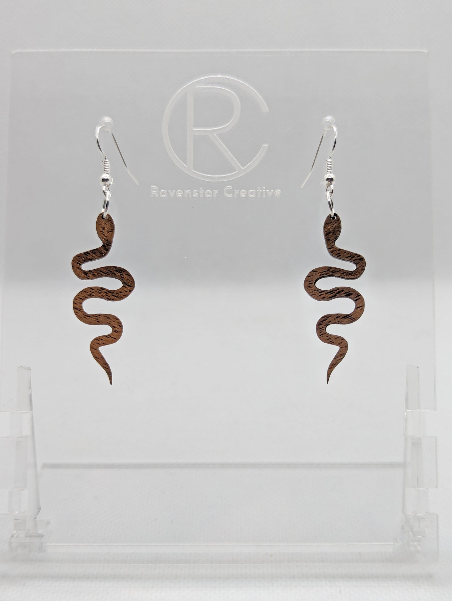 Shows a pair of wooden earrings with silver ear wire hook fixings. They are snake shapes. The colour is Walnut Wood. They are against a white background on a clear stand with the Ravenstor Creative logo on it.