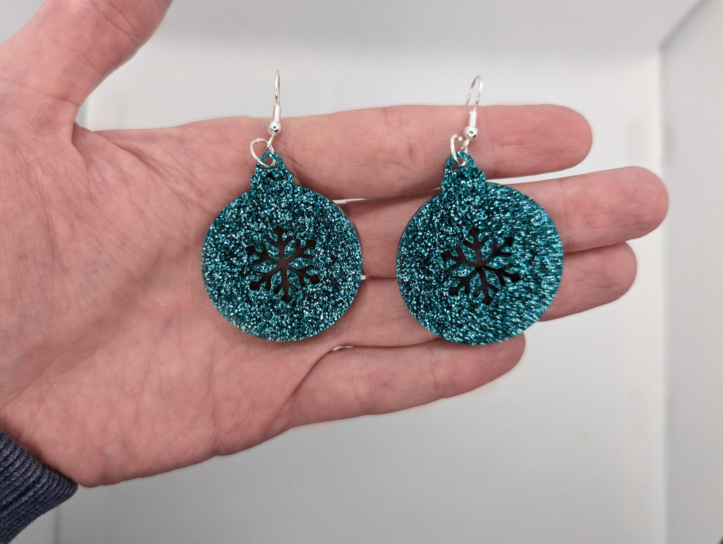 Image of acrylic earrings. They are Christmas bauble shapes with a snowflake cut out in Ice Blue Glitter on a hand showing the size against a white background, they are 2 finger widths length the same again wide. They also have silver fixings.