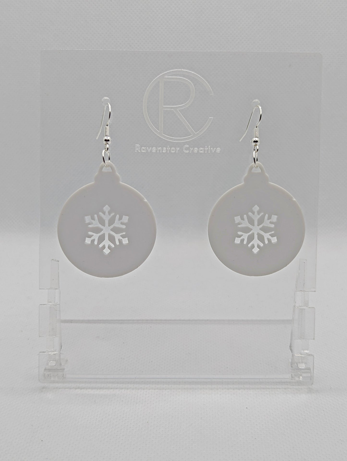 Shows a pair of acrylic earrings that have silver ear wire hook fixings. They are Christmas / Xmas baubles shapes with a snowflake shape cut out the middle. The colour is White. They are against a white background on see through stands.