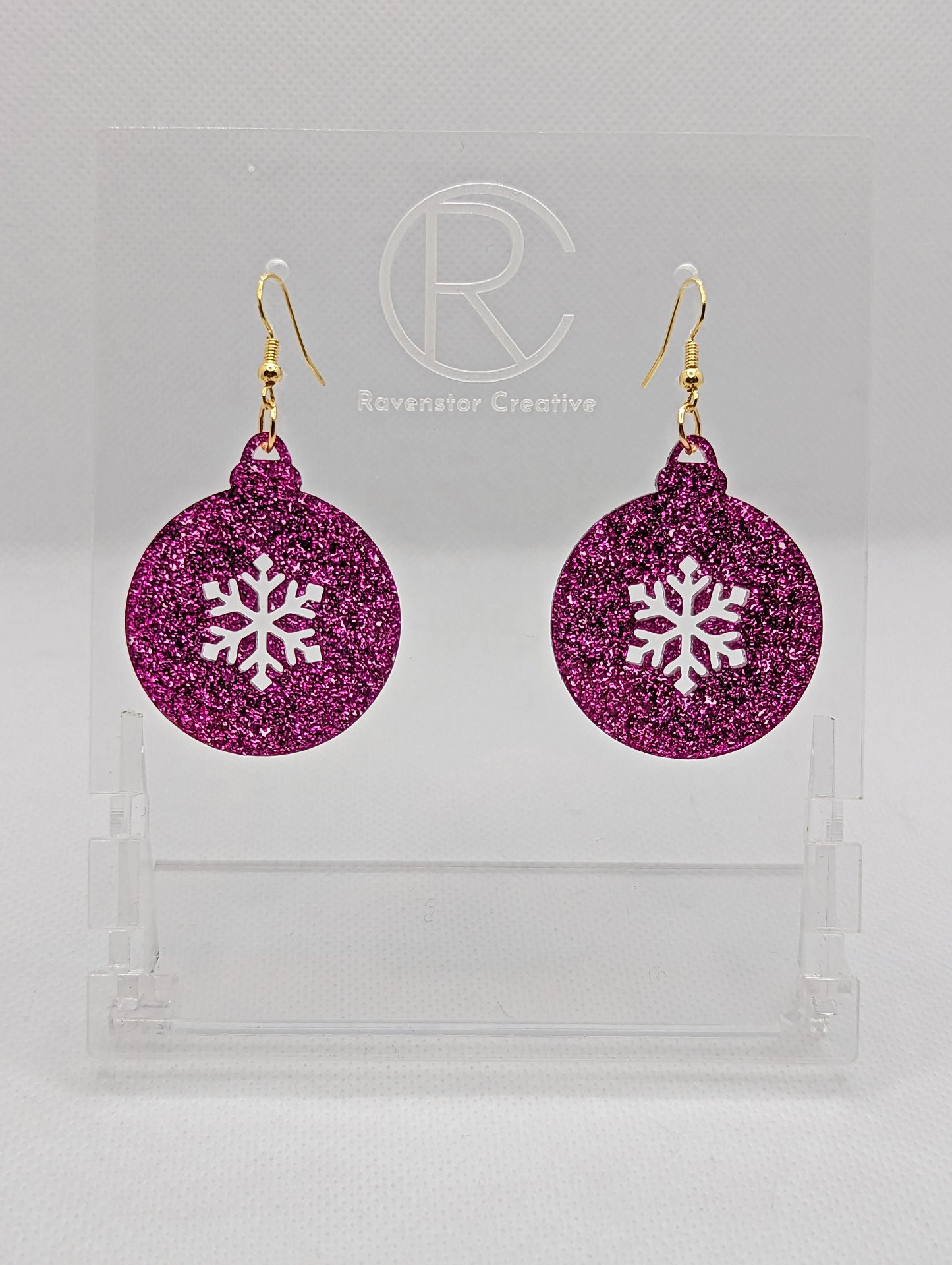Shows a pair of acrylic earrings that have gold ear wire hook fixings. They are Christmas / Xmas baubles shapes with a snowflake shape cut out the middle. The colour is Pink Glitter. They are against a white background on see through stands.