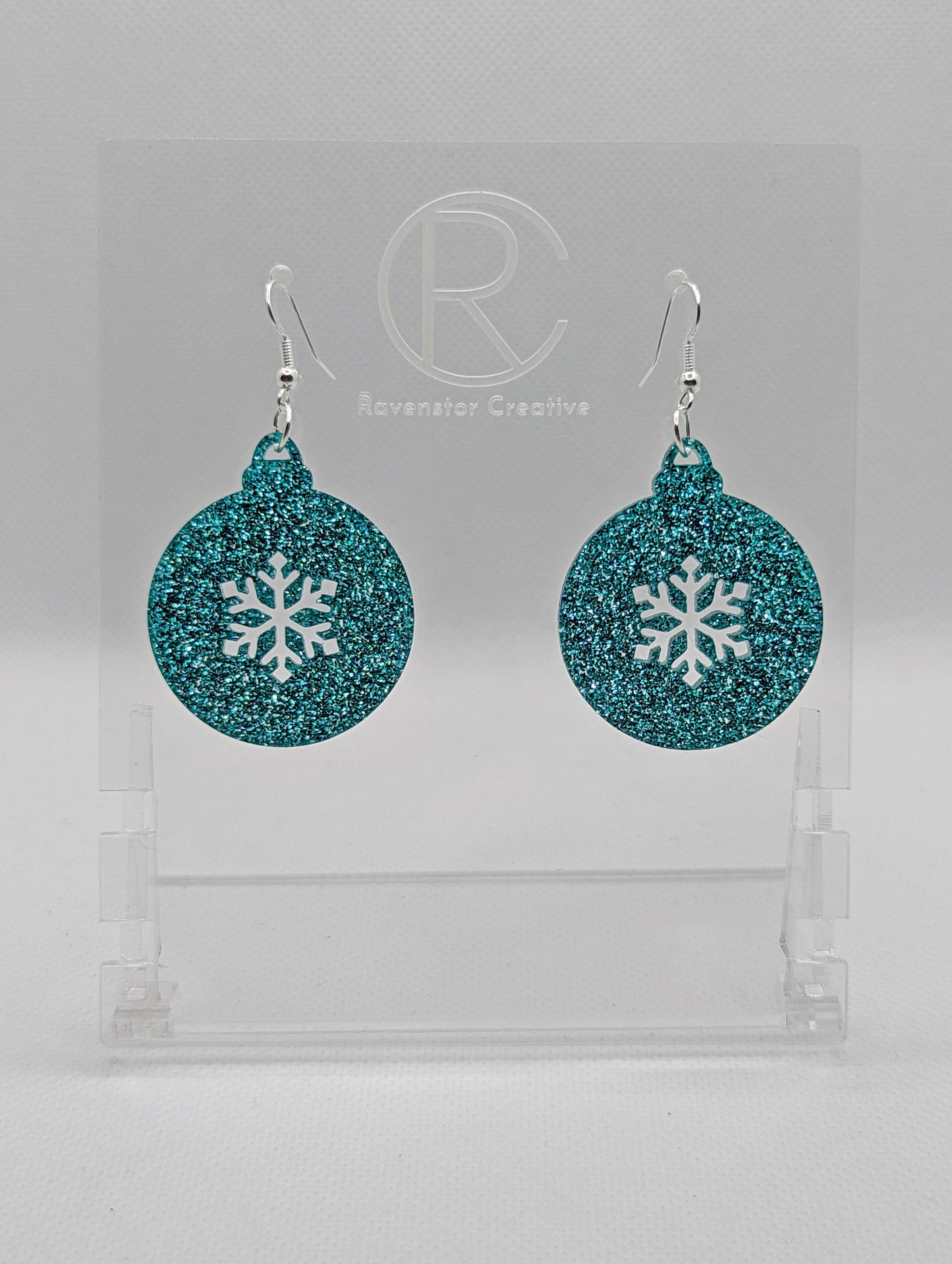 Shows a pair of acrylic earrings that have silver ear wire hook fixings. They are Christmas / Xmas baubles shapes with a snowflake shape cut out the middle. The colour is Ice Blue Glitter. They are against a white background on see through stands.
