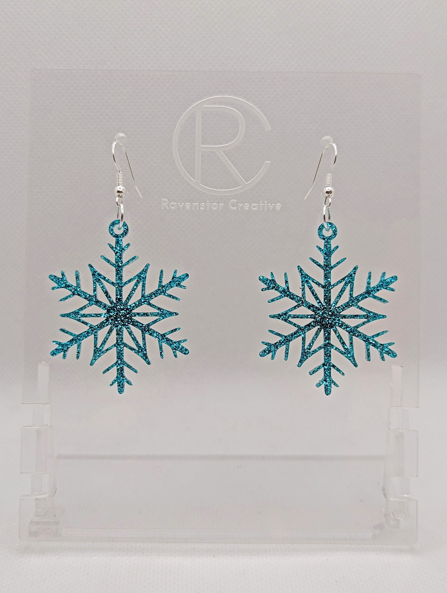 A pair of earrings that have silver ear wire hook fixings. They are elegant snowflake shapes. They are ice blue glitter in colour and are against a white background on see through stand with the Ravenstor Creative logo on it. Perfect for Christmas