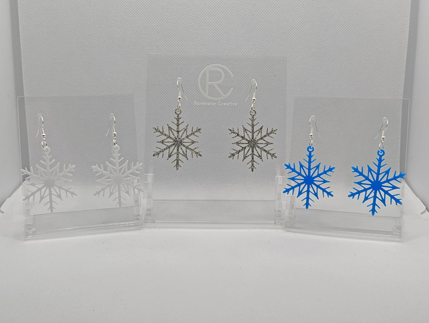 Shows three pairs of earrings that have silver ear wire hook fixings. They are elegant snowflake shapes. The colours are white, silver glitter and blue. They are against a white background on see through stand with the ravenstor creative logo on.