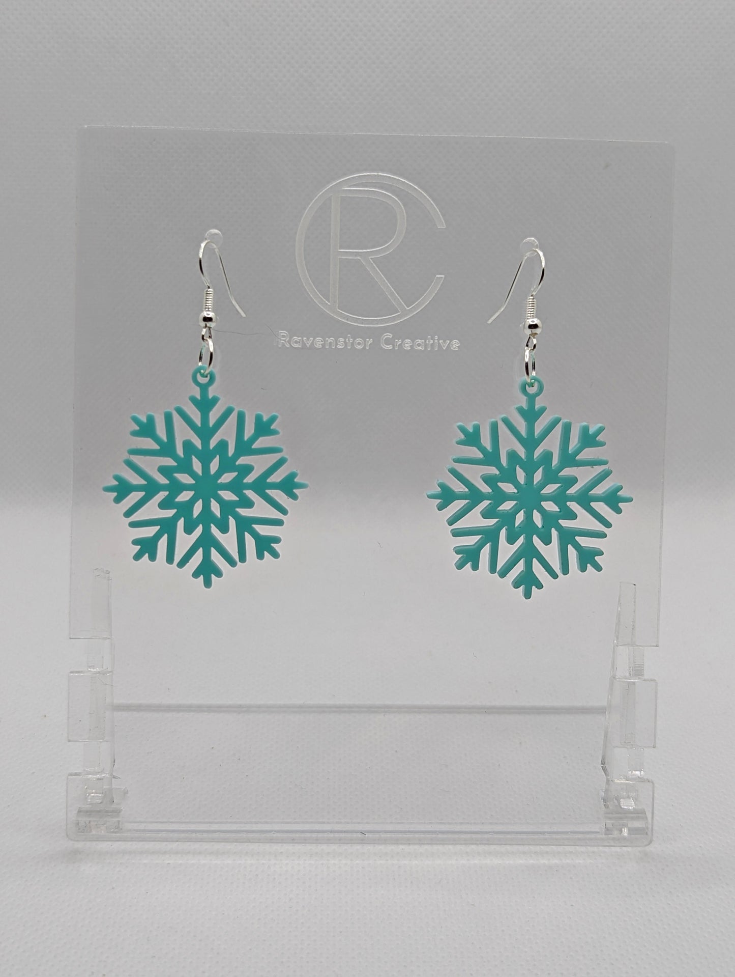A pair of earrings that have silver ear wire hook fixings. They are elegant snowflake shapes. They are spearmint in colour and are against a white background on see through stand with the Ravenstor Creative logo on it. Perfect for Christmas