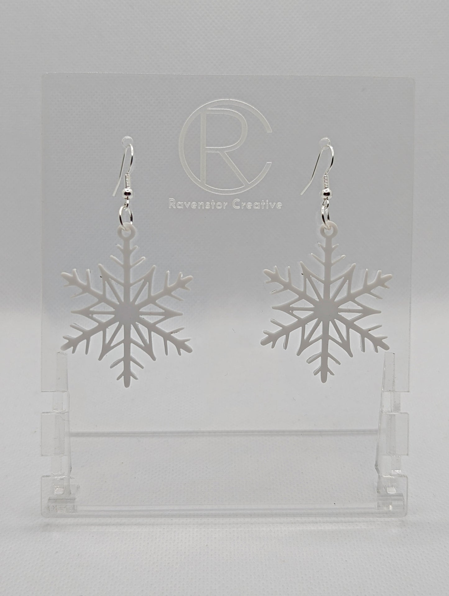 A pair of earrings that have silver ear wire hook fixings. They are elegant snowflake shapes. They are white in colour and are against a white background on see through stand with the Ravenstor Creative logo on it. Perfect for Christmas