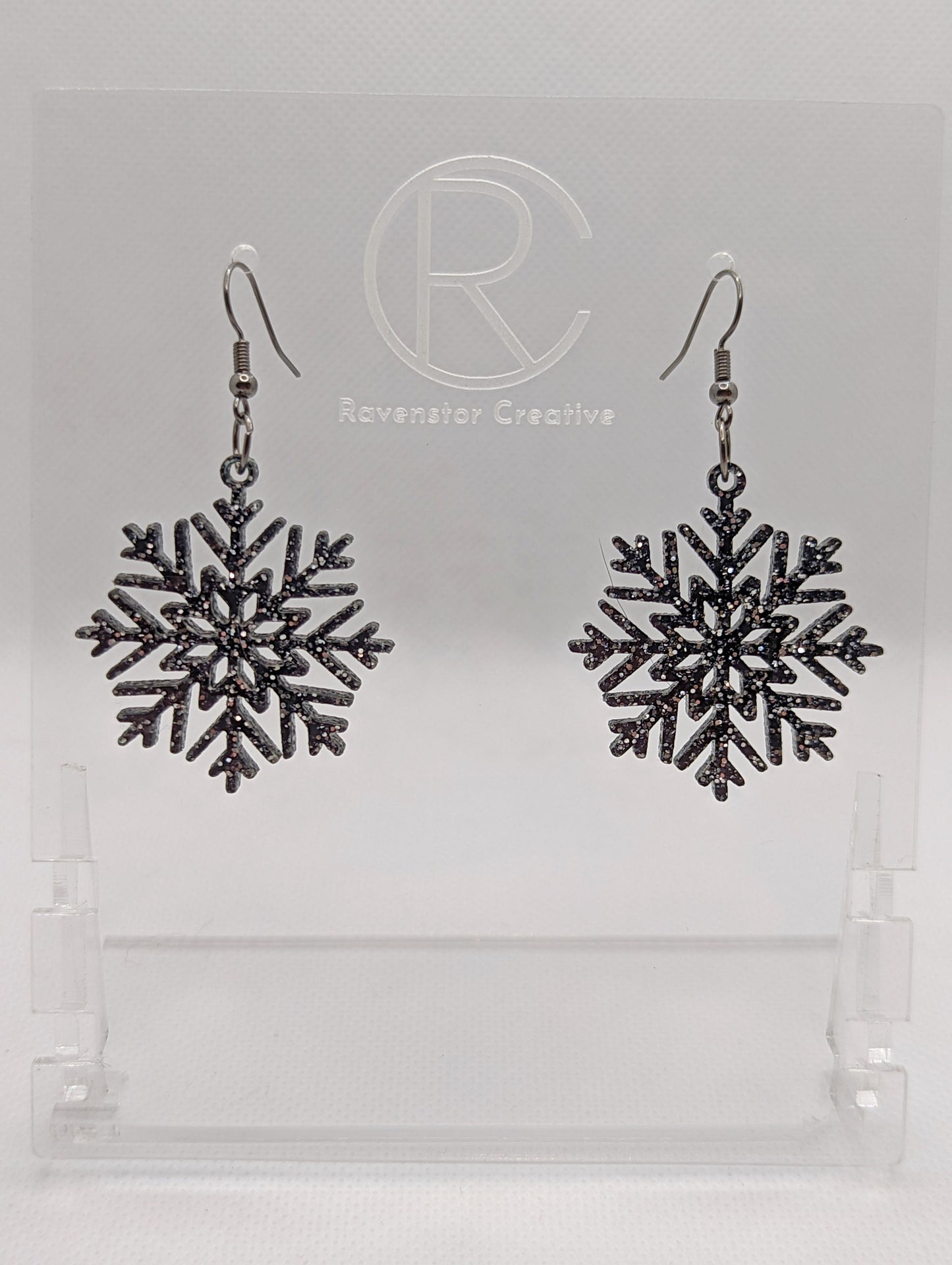 A pair of earrings that have black ear wire hook fixings. They are elegant snowflake shapes. They are Black & Silver glitter in colour and are against a white background on see through stand with the Ravenstor Creative logo on it. Perfect for Winter