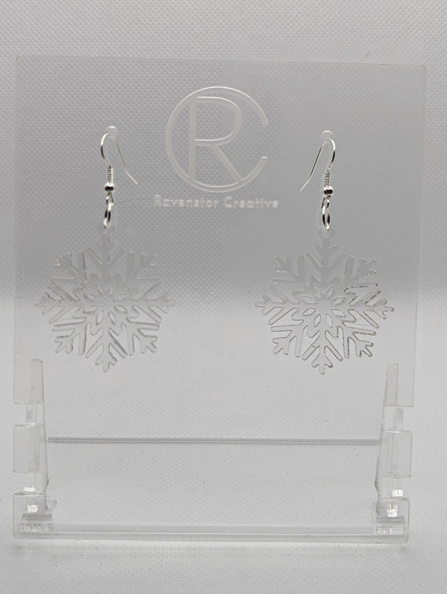 A pair of earrings that have silver ear wire hook fixings. They are elegant snowflake shapes. They are clear / See through in colour and are against a white background on see through stand with the Ravenstor Creative logo on it. Perfect for Christmas