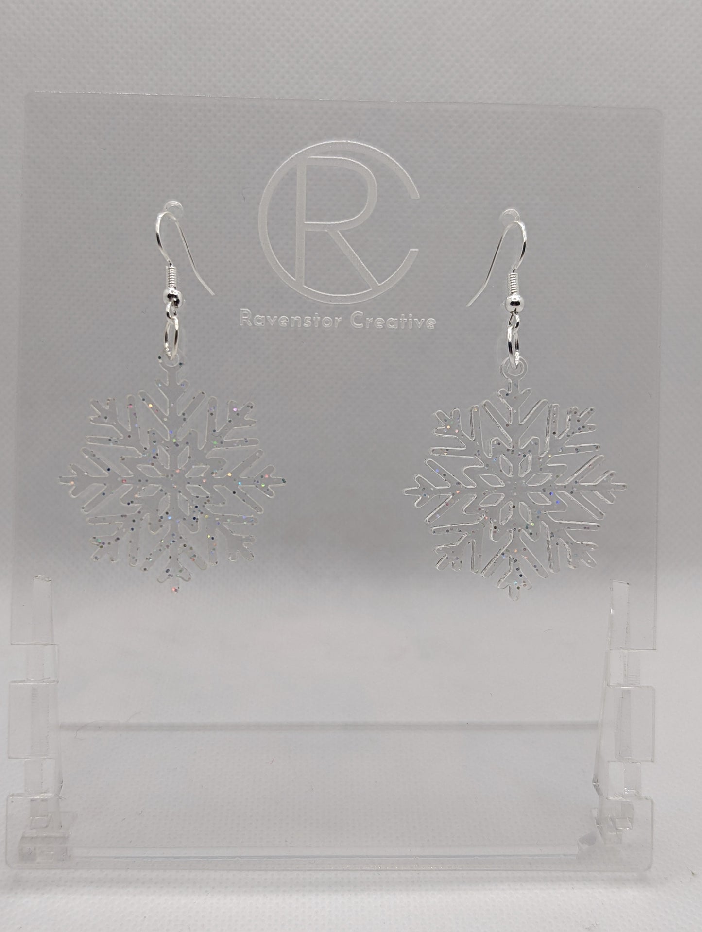 A pair of earrings that have silver ear wire hook fixings. They are elegant snowflake shapes. They are clear sparkle glitter in colour and are against a white background on see through stand with the Ravenstor Creative logo on it. Perfect for xmas