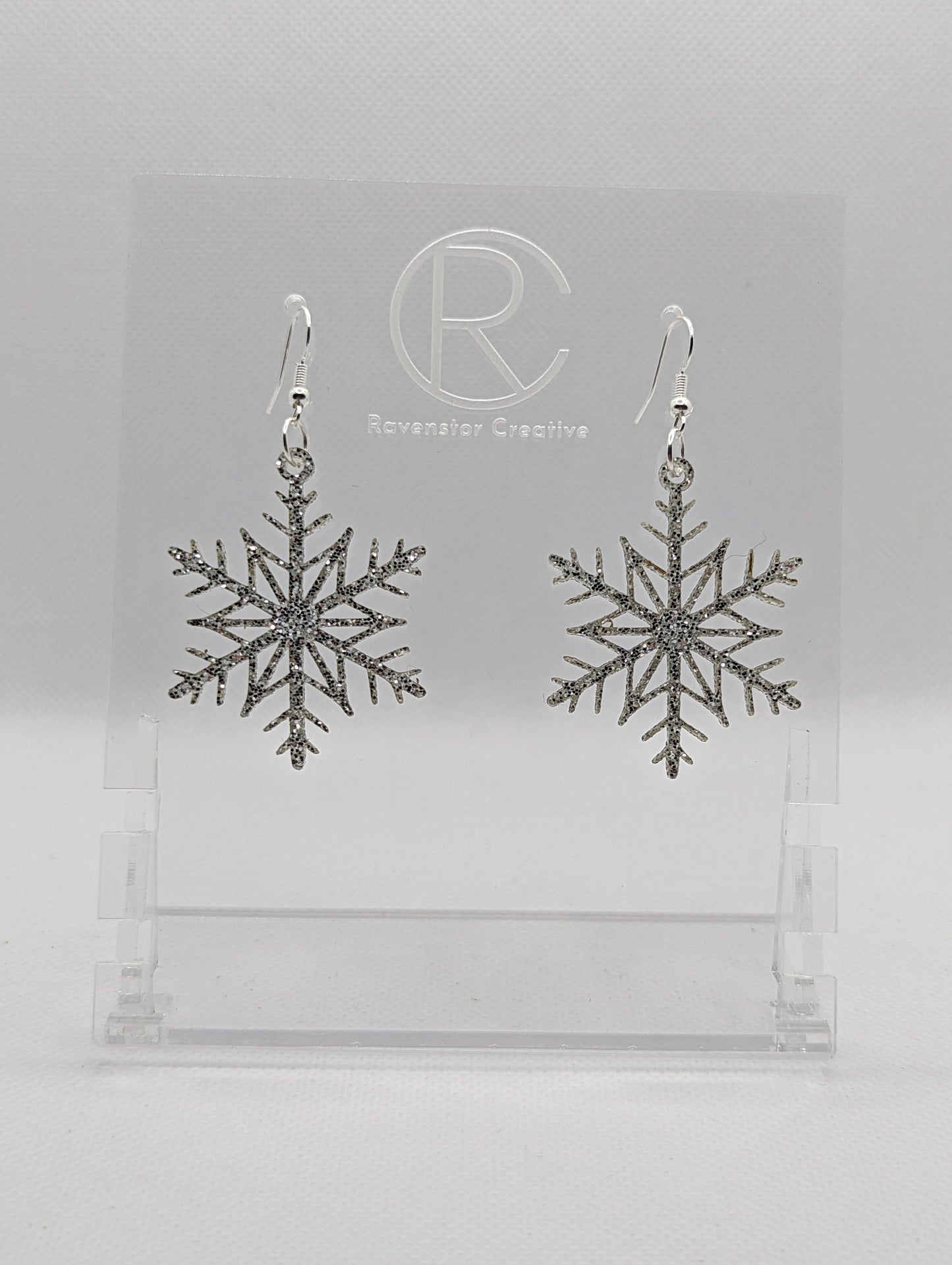 A pair of earrings that have silver ear wire hook fixings. They are elegant snowflake shapes. They are Silver glitter in colour and are against a white background on see through stand with the Ravenstor Creative logo on it. Perfect for Winter