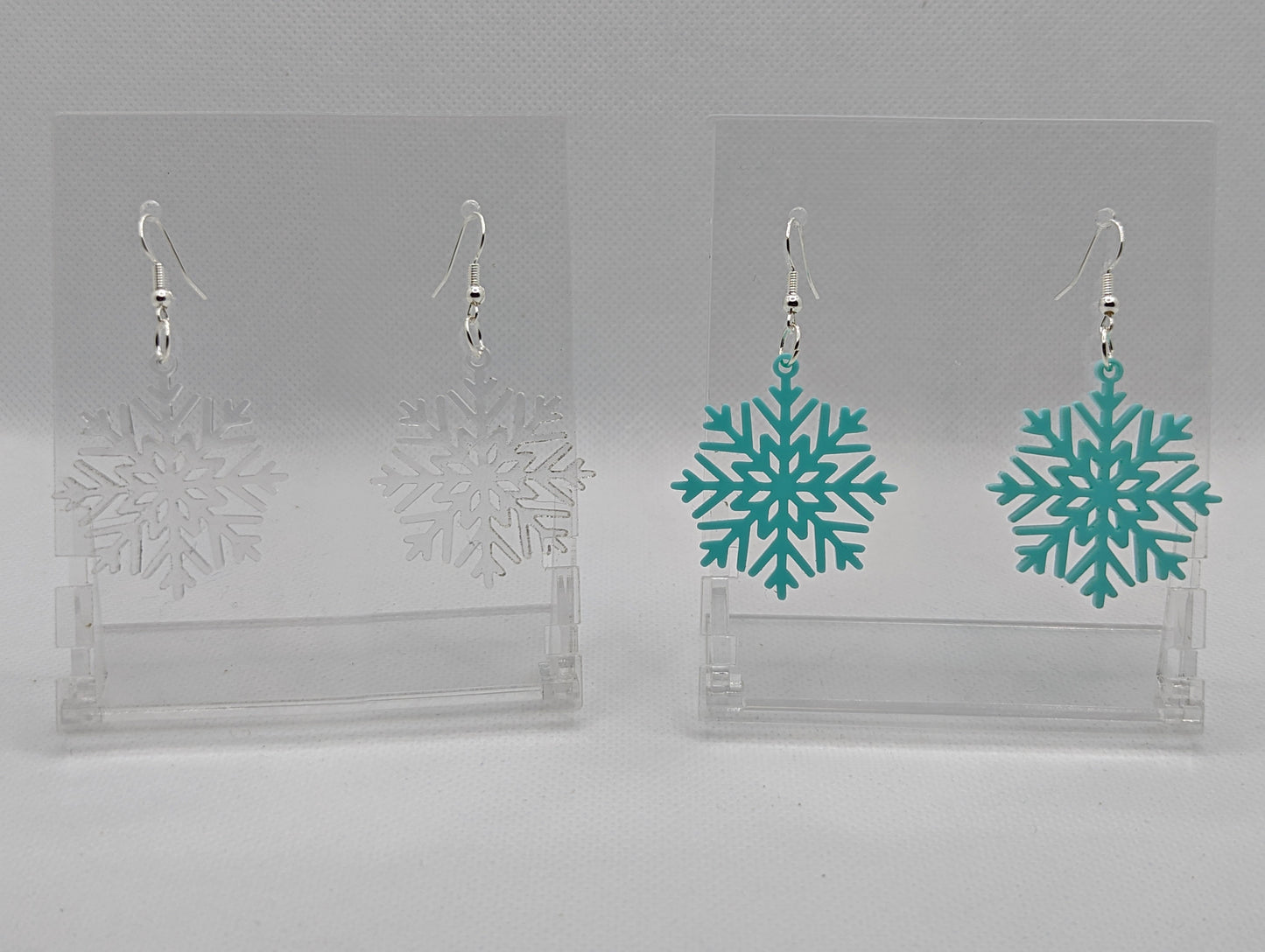 Shows two pairs of earrings that have silver ear wire hook fixings. They are elegant snowflake shapes. The colours are clear / See through and spearmint. They are against a white background on see through stand with the ravenstor creative logo on it.