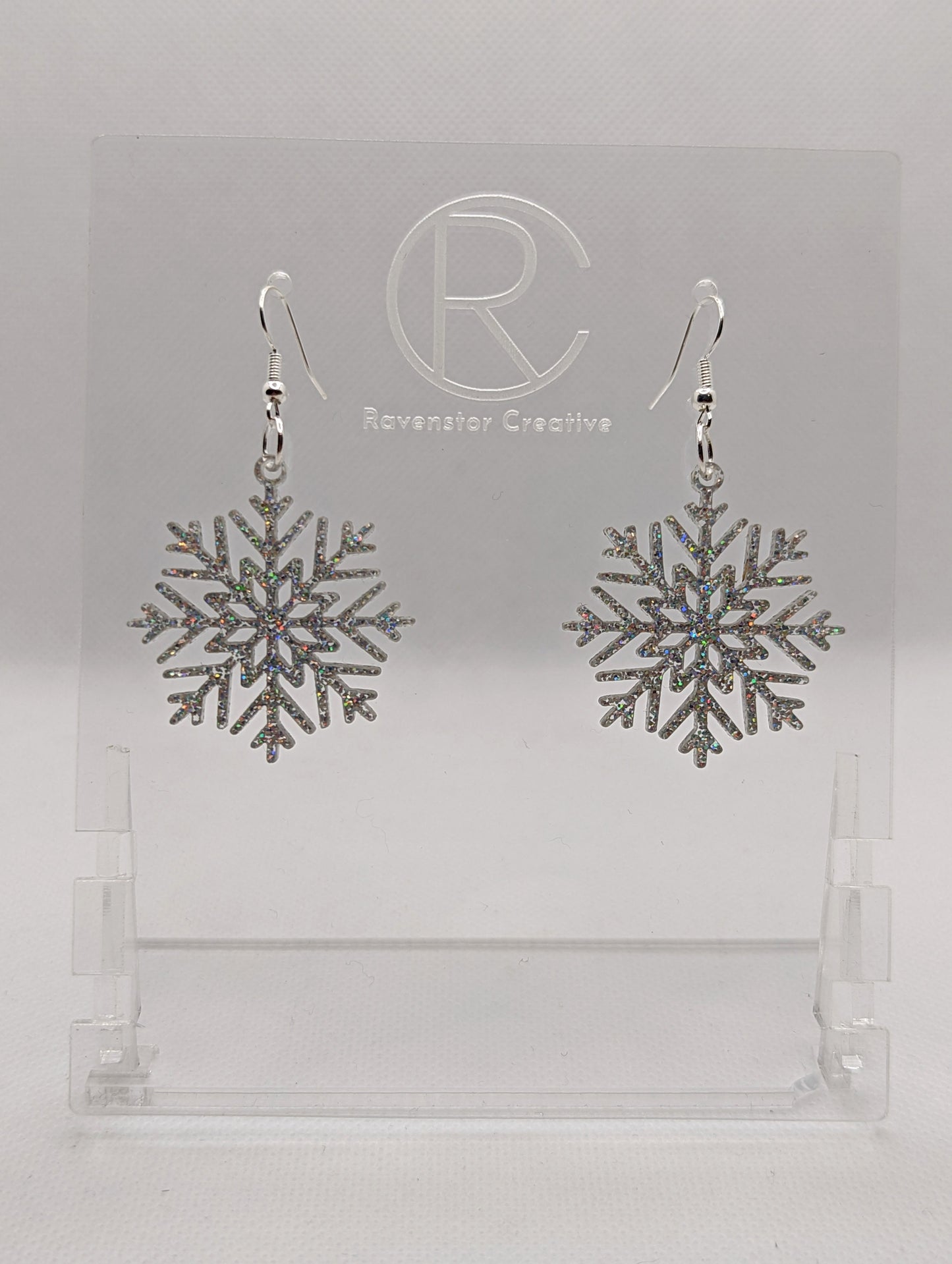 A pair of earrings that have silver ear wire hook fixings. They are elegant snowflake shapes. They are holographic glitter in colour and are against a white background on see through stand with the Ravenstor Creative logo on it. Perfect for Christmas