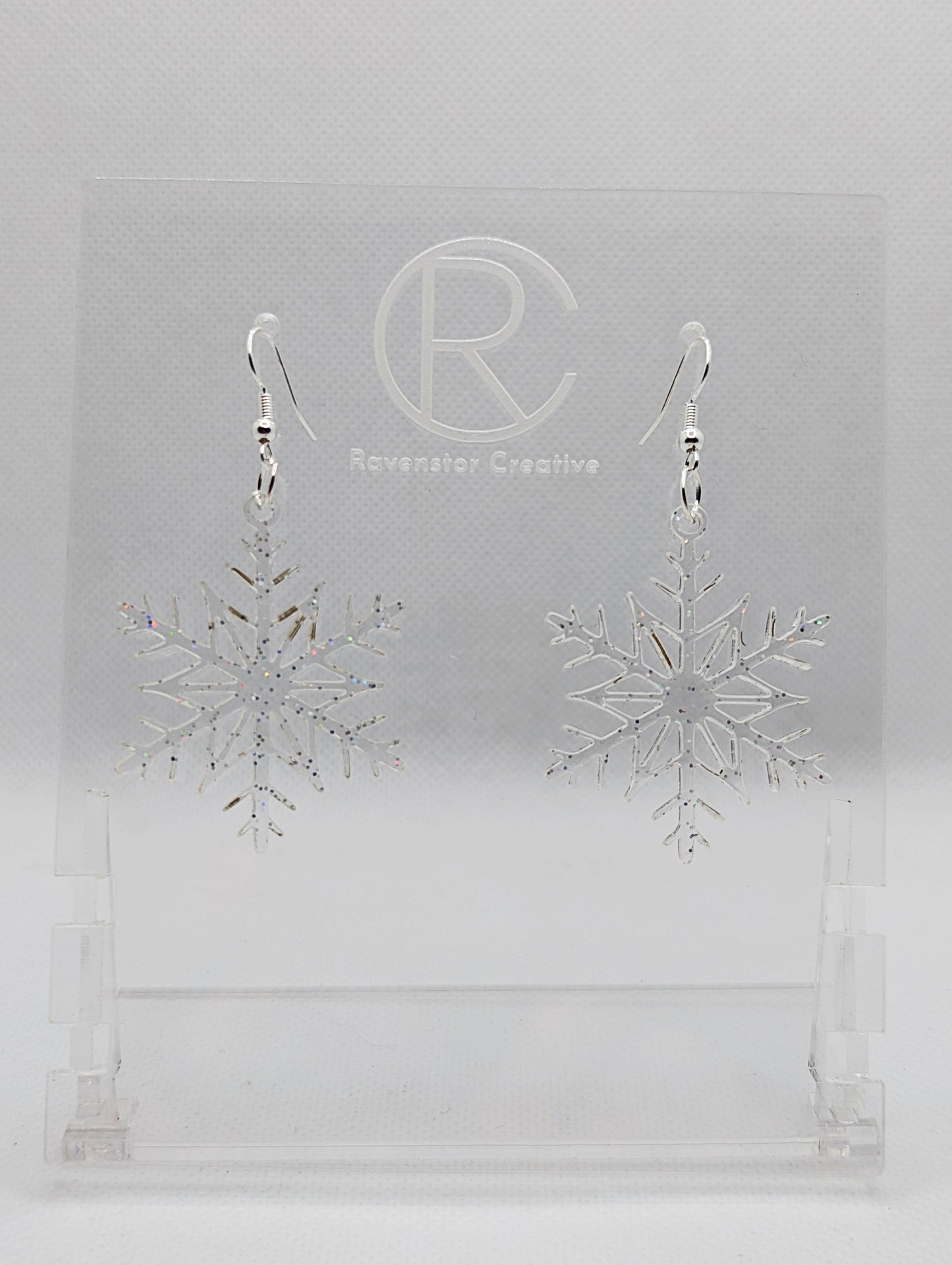 A pair of earrings that have silver ear wire hook fixings. They are elegant snowflake shapes. They are clear sparkle glitter in colour and are against a white background on see through stand with the Ravenstor Creative logo on it. Perfect for Xmas.