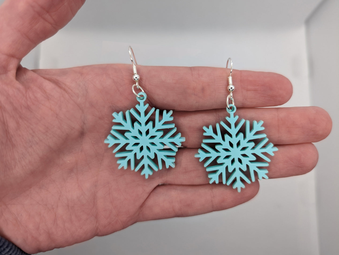 Image of a pair of elegant snowflake shaped earrings on a hand showing the size against a white background, they are two and a half finger widths length and same again wide. They are spearmint in colour and have silver fixings. Perfect for Christmas