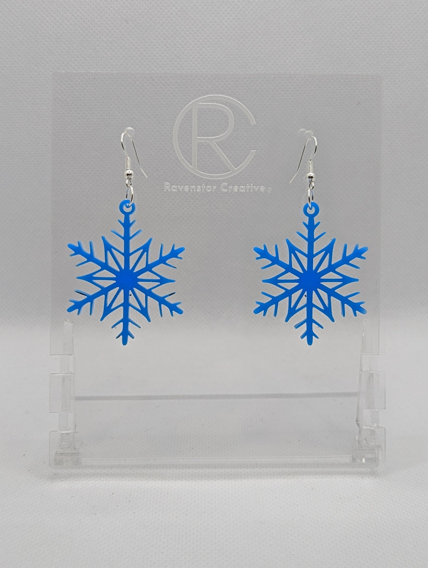 A pair of earrings that have silver ear wire hook fixings. They are elegant snowflake shapes. They are blue in colour and are against a white background on see through stand with the Ravenstor Creative logo on it. Perfect for Christmas