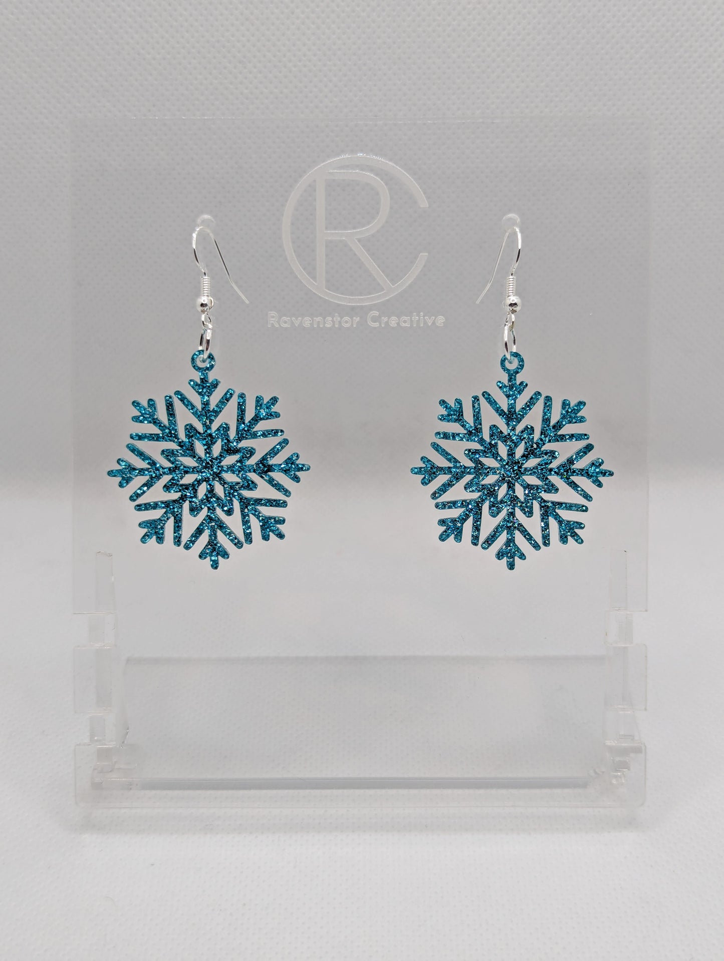 A pair of earrings that have silver ear wire hook fixings. They are elegant snowflake shapes. They are Ice Blue glitter in colour and are against a white background on see through stand with the Ravenstor Creative logo on it. Perfect for Christmas